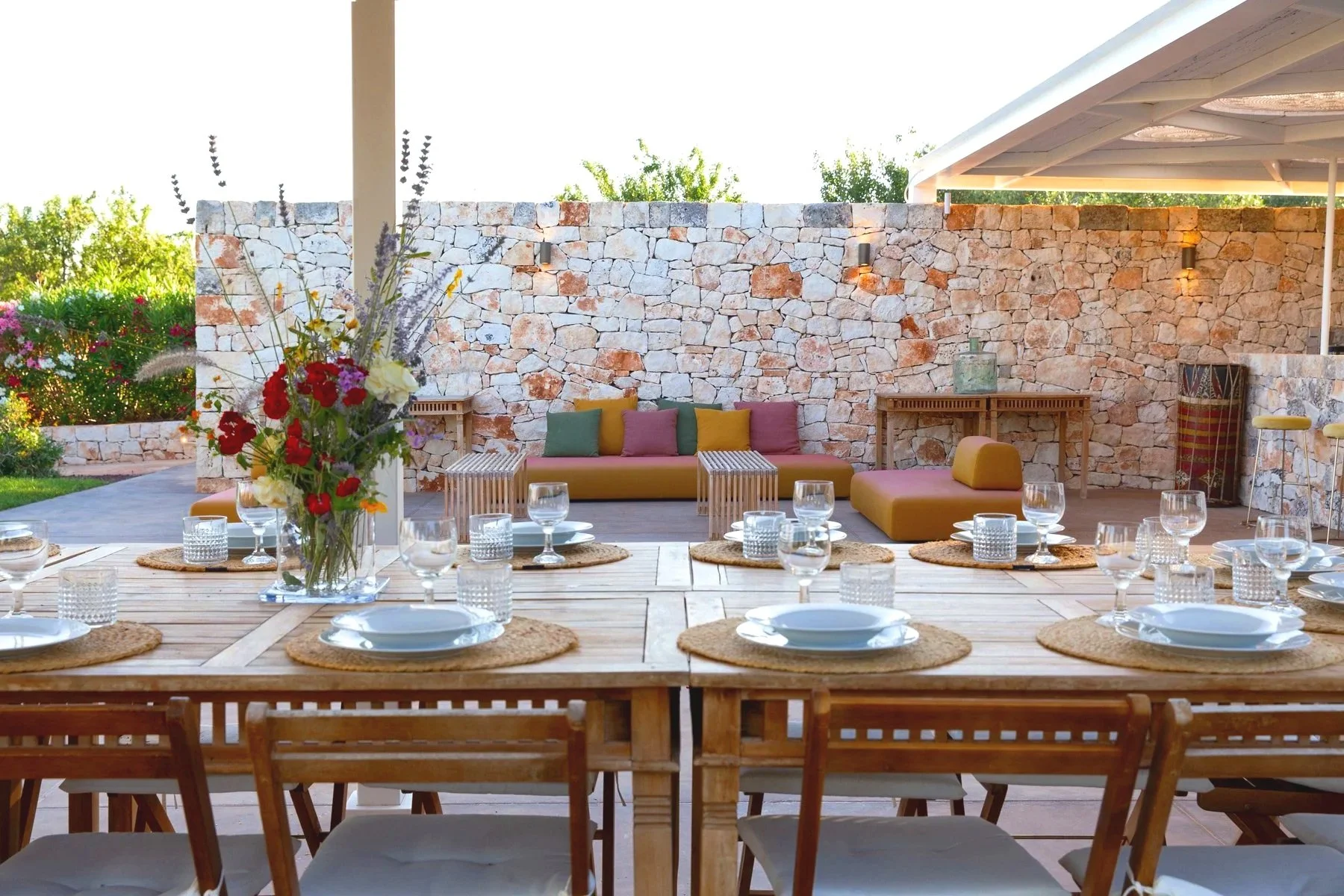 Espace extérieur avec une grande table en bois, décorée de verres à vin, assiettes et sets de table en raphia, au centre une vase avec des fleurs colorées, en arrière-plan un mur en pierre avec un banc en tissu jaune et coussins multicolores, une con