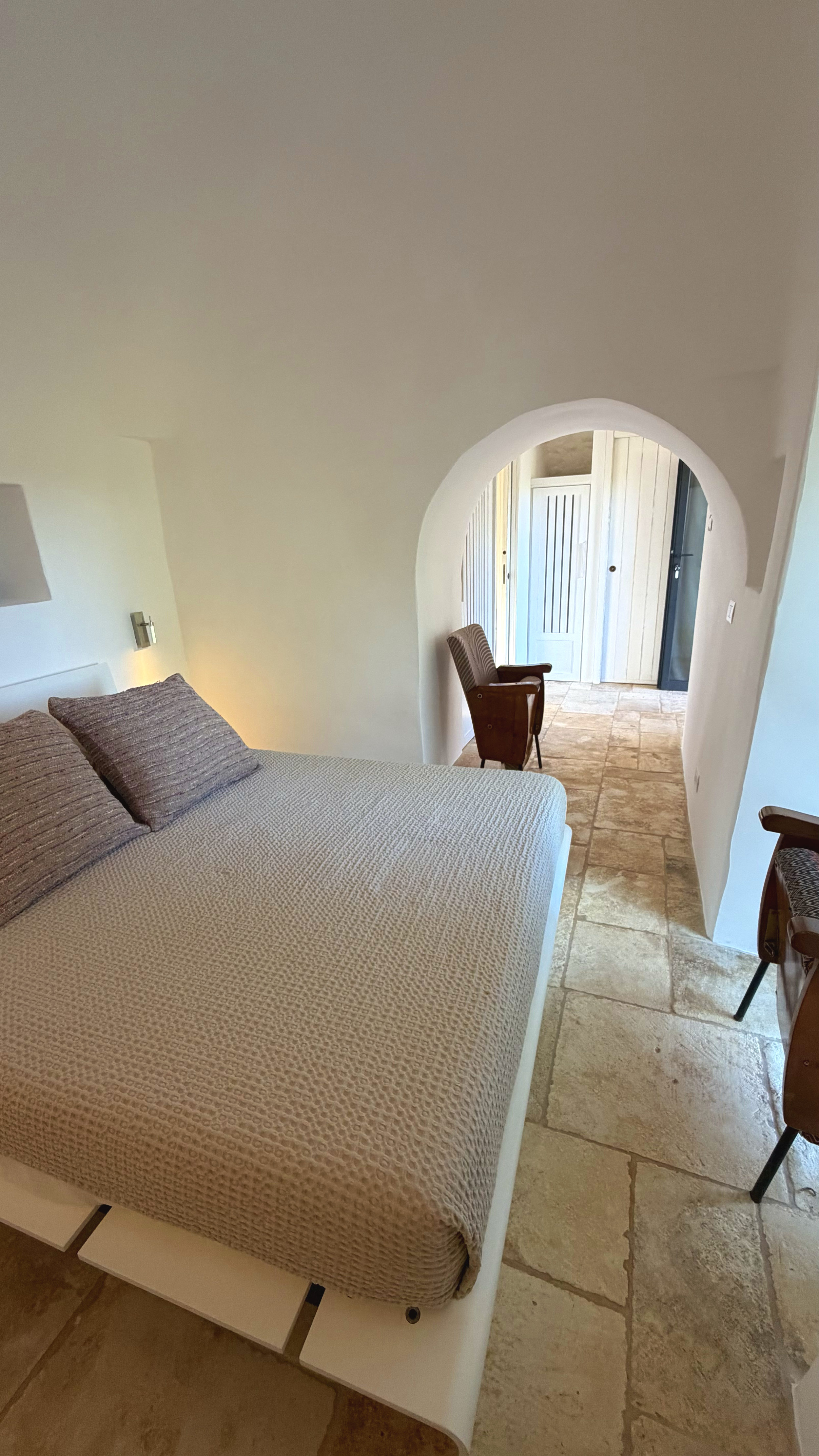 Une chambre d'hôtel avec un lit, un fauteuil en bois, une arche menant à une entrée lumineuse et un sol en pierre.