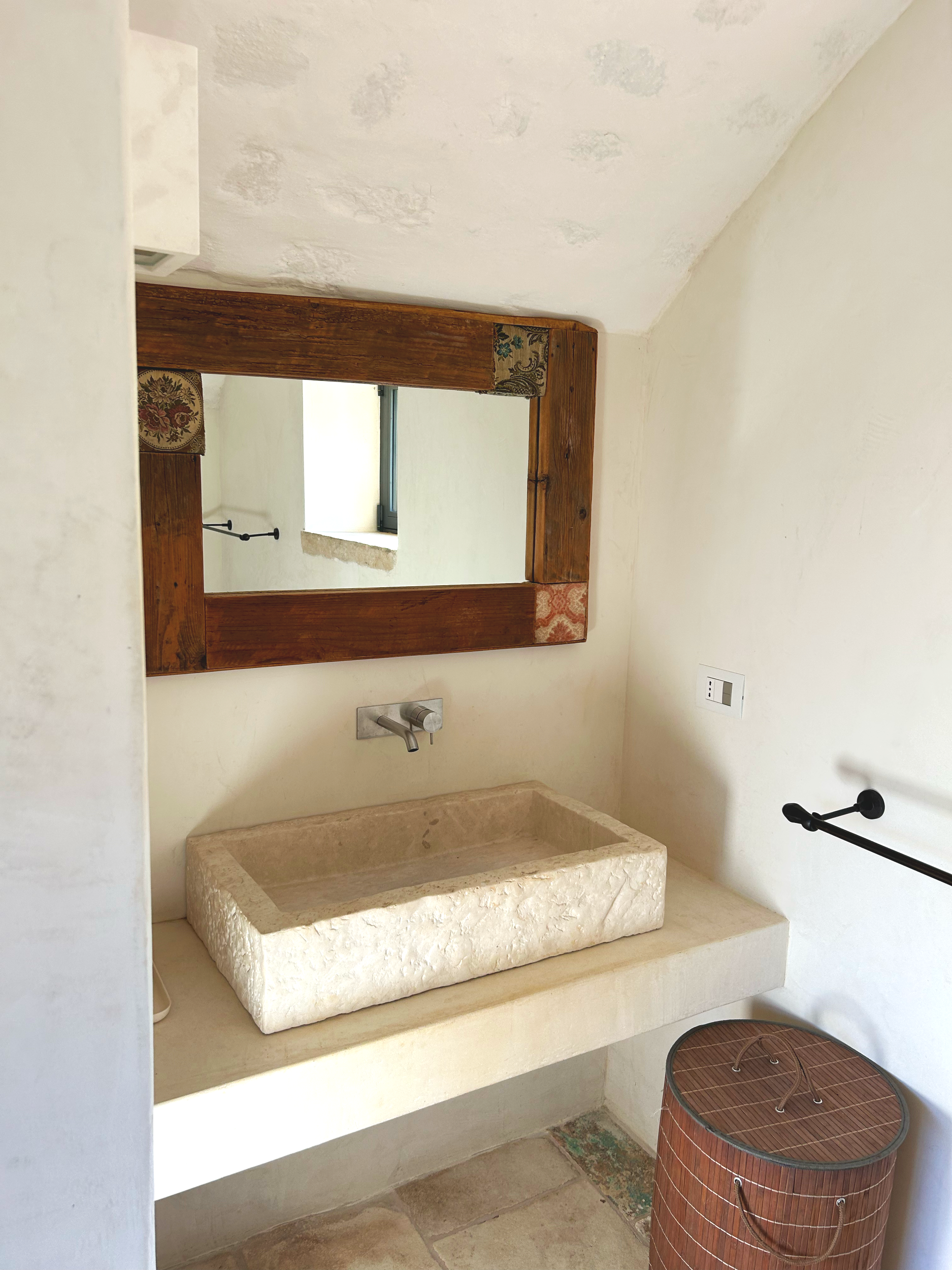 Un miroir encadré en bois au-dessus d'un lavabo en pierre, d'une petite fenêtre, d'un robinet, d'une serviette noire accrochée à un support, et d'une boîte de rangement en bois.