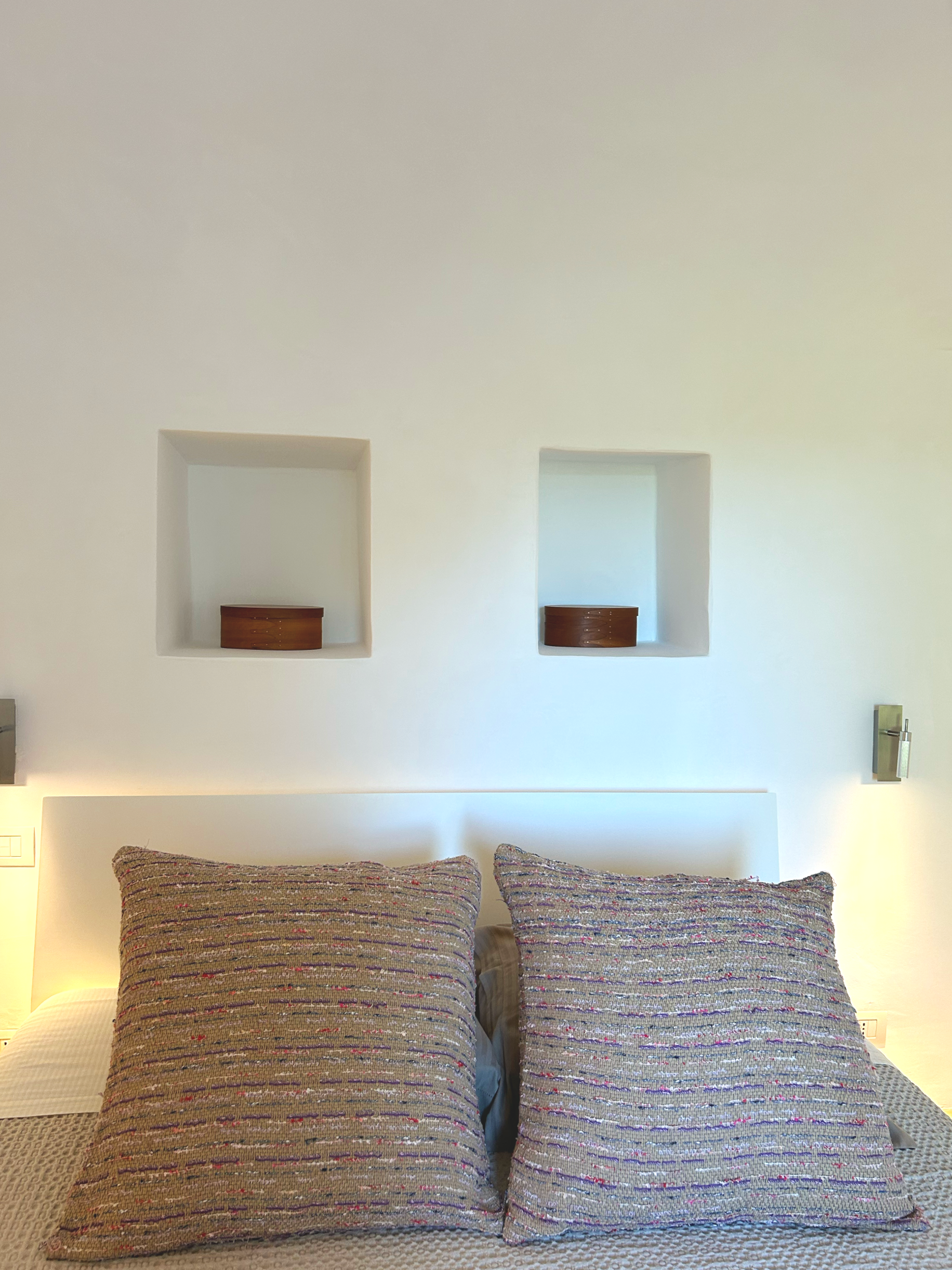 Deux oreillers rayés posés sur un lit, avec des lampes de chaque côté et deux niches murales au-dessus, contenant des objets en bois.