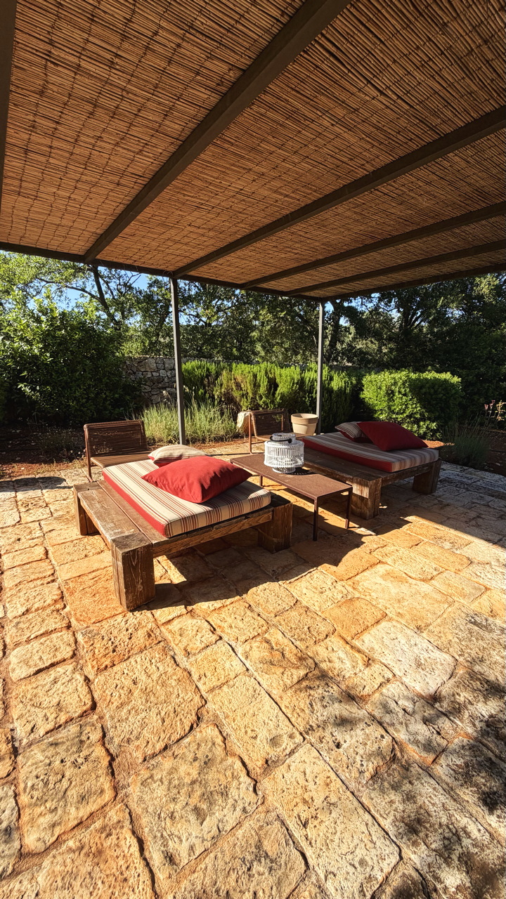 Deux lits de jour en bois avec coussins rouges sous un toit en canne, sur une terrasse en pierre, entourée de végétation et d'arbres.