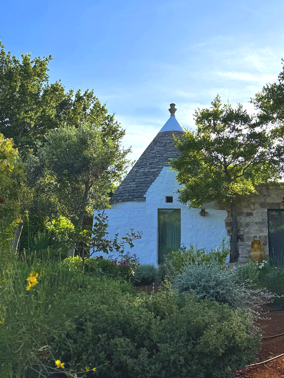 Une petite maison en pierre avec un toit conique en tuiles, entourée d'arbres et de plantes dans un jardin en plein air, sous un ciel bleu.