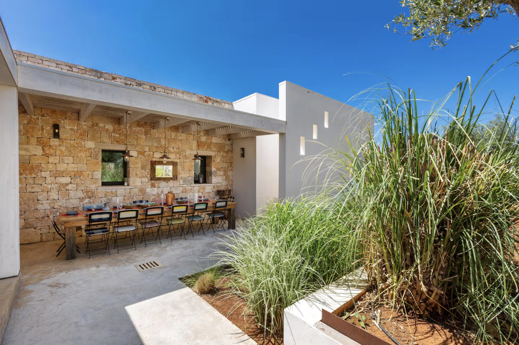 Une terrasse extérieure attenante à une maison moderne, avec un mur en pierre, une longue table en bois avec des chaises autour, et un jardin avec de hautes herbes et des plantes, sous un ciel bleu clair.