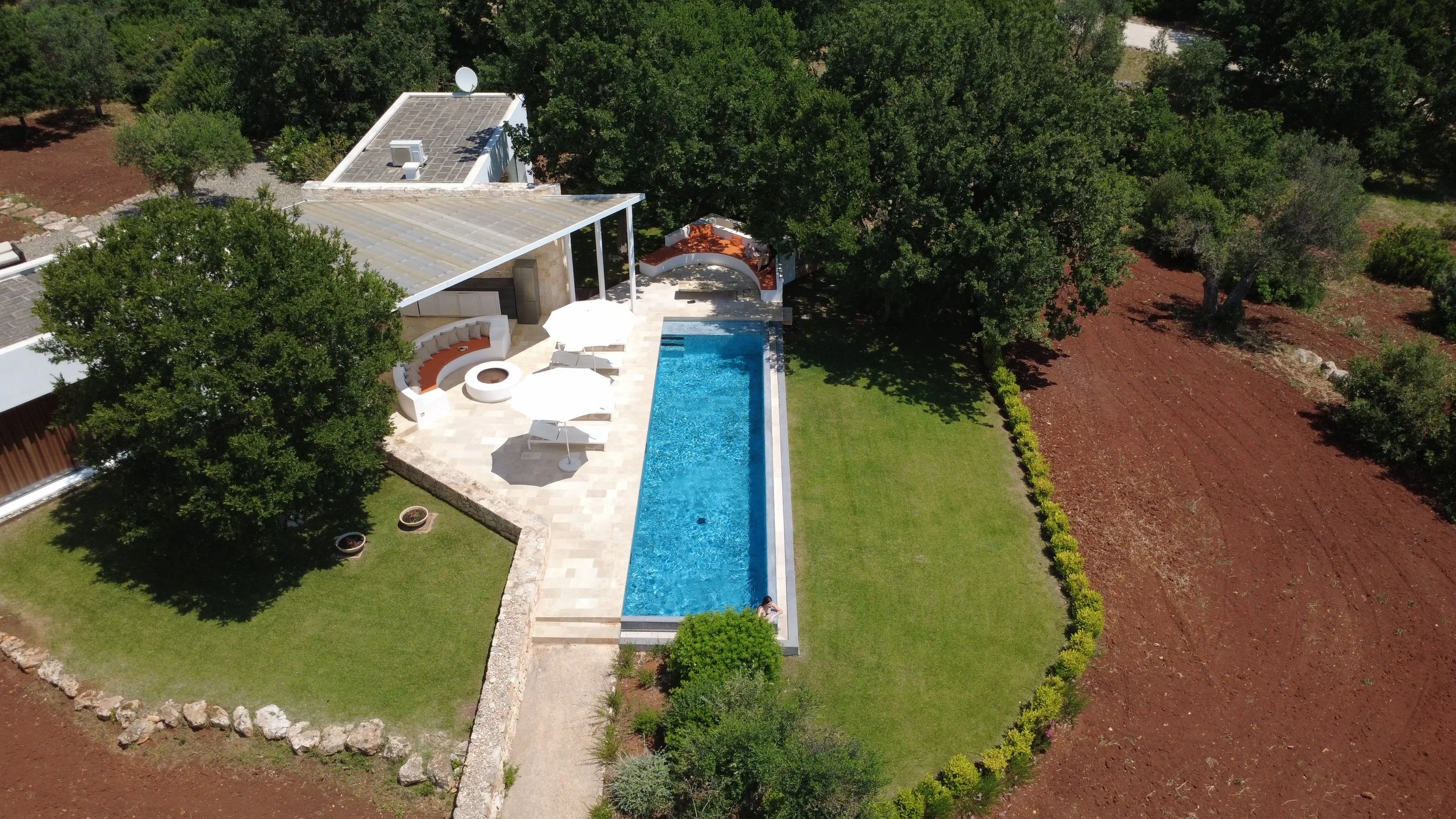 Vue aérienne d'une piscine rectangulaire avec une zone de détente avec parasols, entourée de pelouse et d'une terrasse, avec des arbres et des champs à proximité.