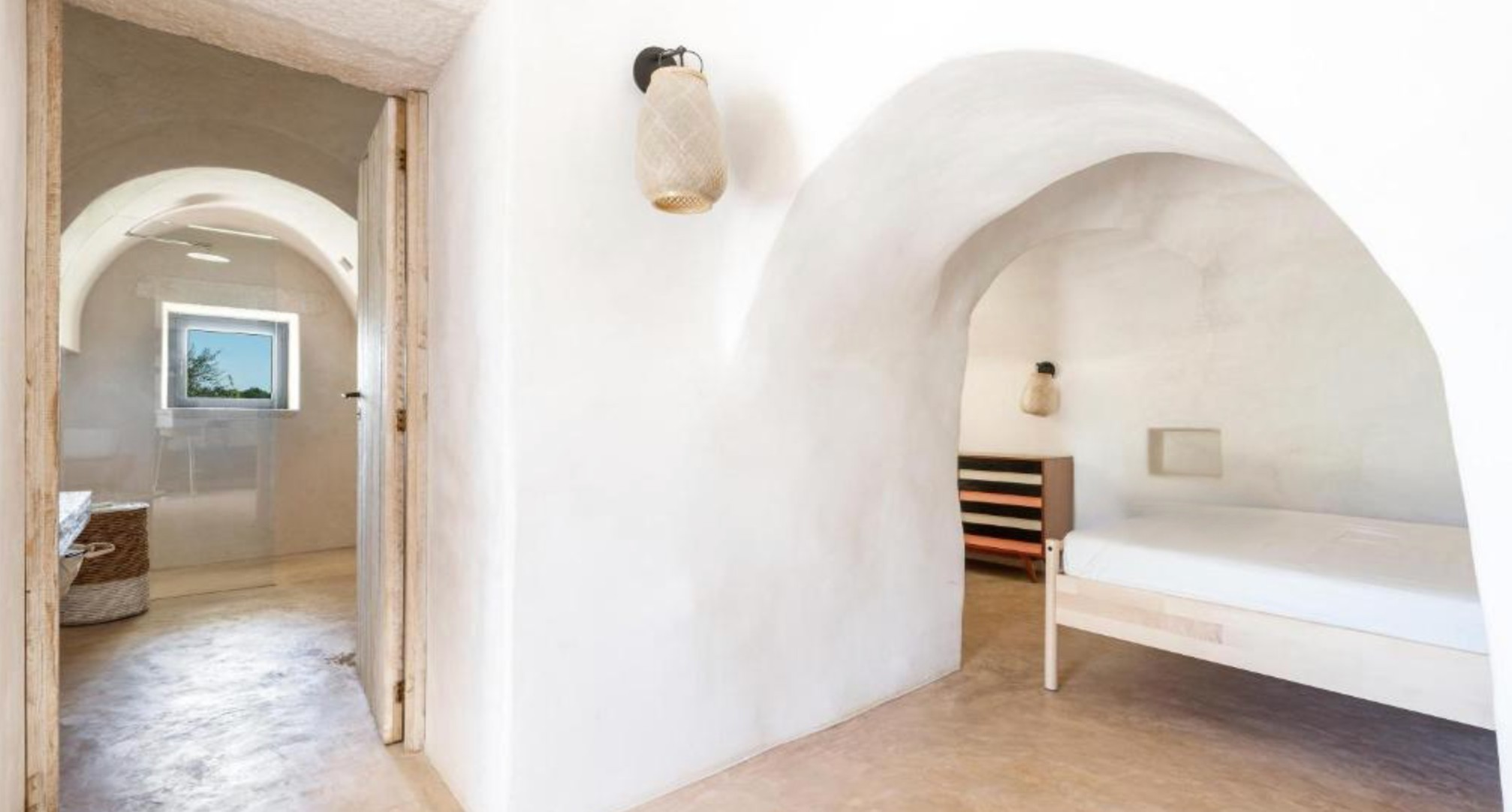 Intérieur d'une maison avec murs et plafonds en forme d'arcs, chambre avec lit simple, lampe suspendue en rotin, et fenêtre offrant une vue sur le ciel et des arbres.