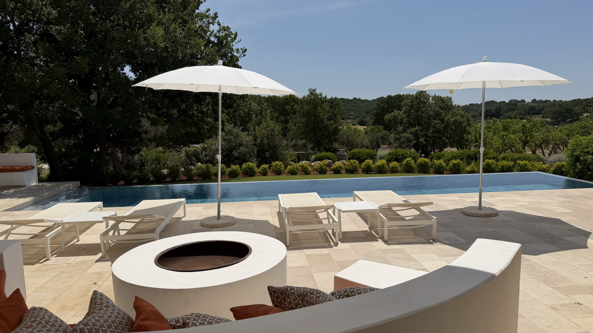 Piscine extérieure avec parasols blancs, chaises longues, vues sur des arbres et un paysage vallonné au loin.