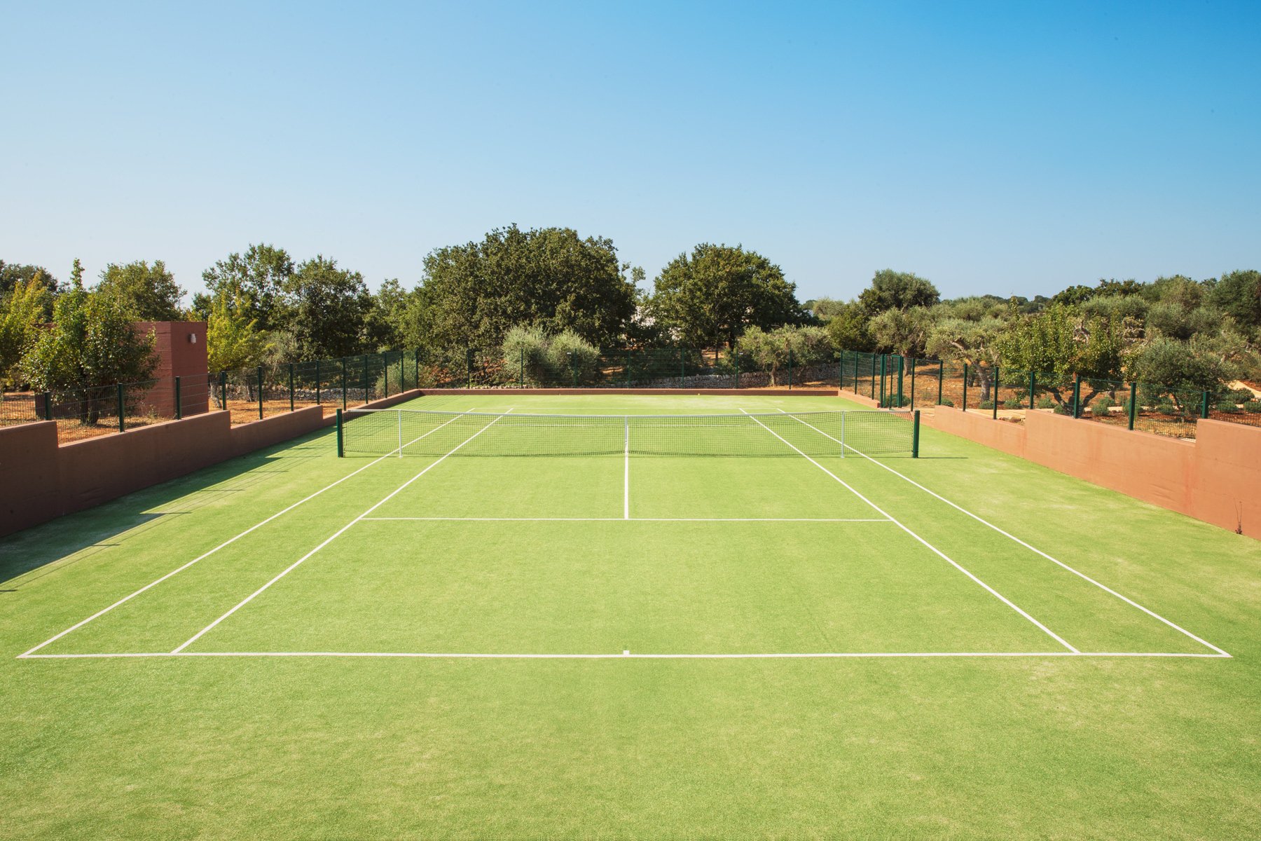Court de tennis en herbe entouré d'un mur en briques et de haies d'arbres sous un ciel clair.