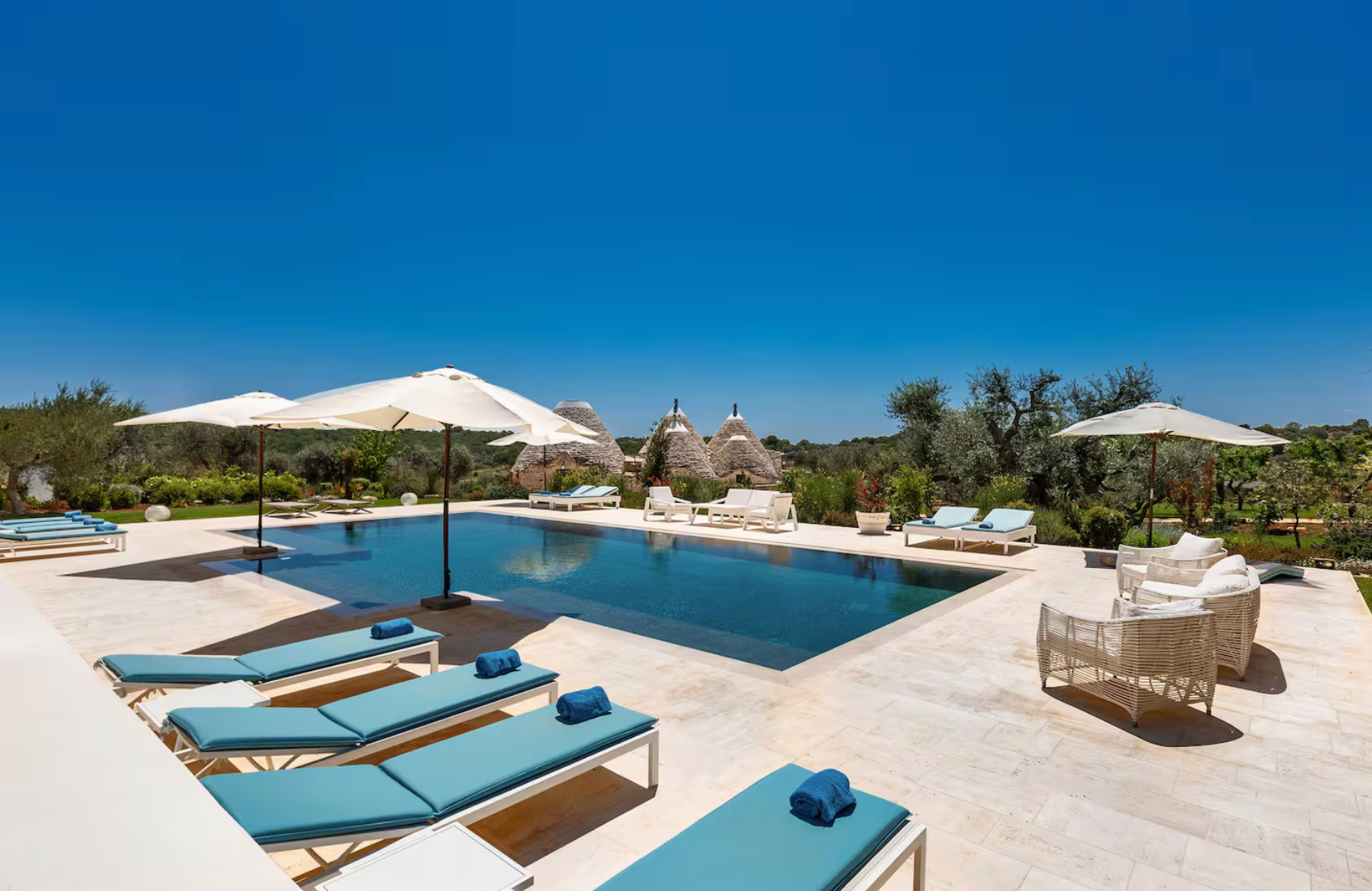 Piscine extérieure avec transats bleus, parasols blancs, chaises en rotin et vue sur des habitations traditionnelles en pierre dans un environnement naturel sous un ciel bleu
