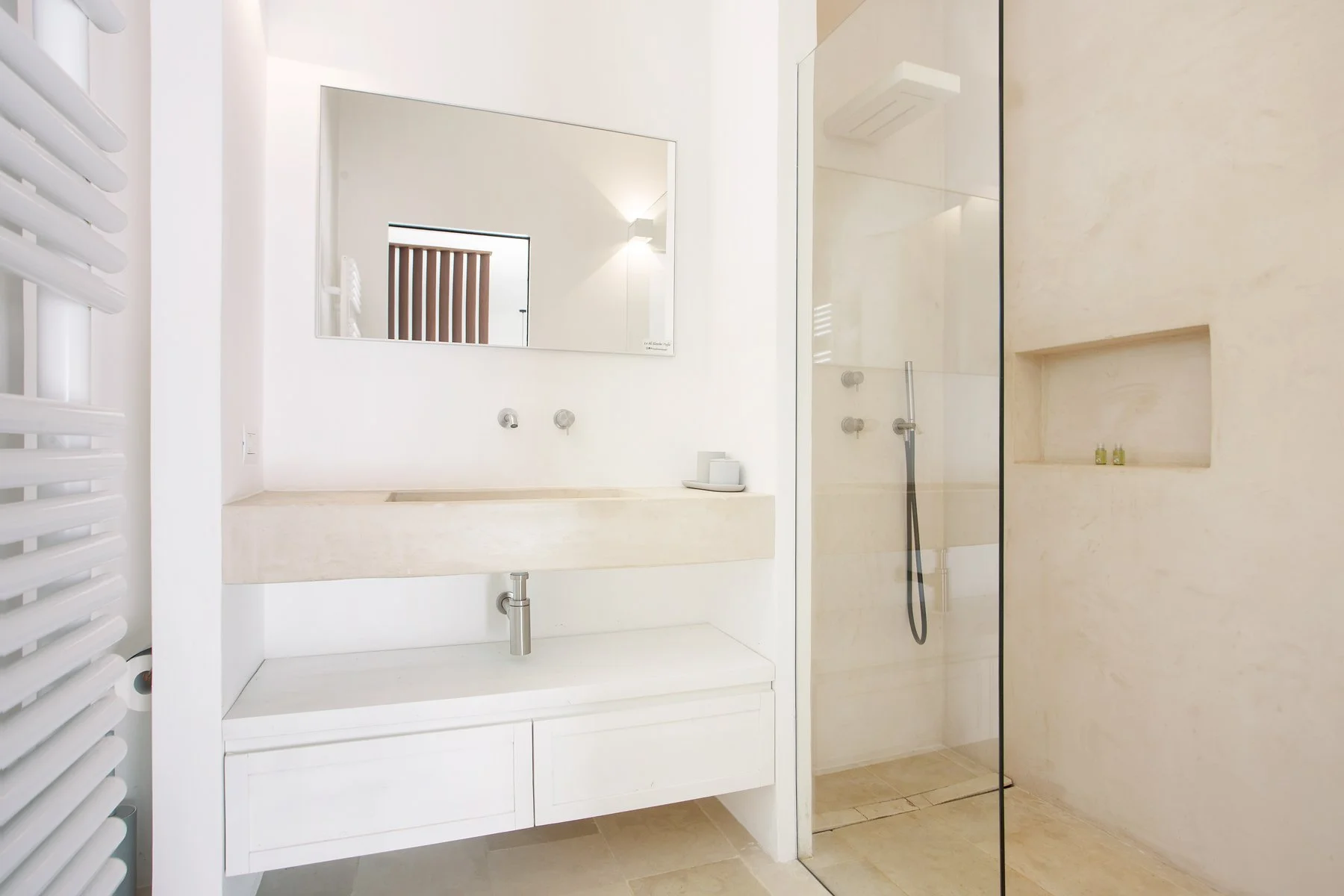 Salle de bains moderne avec lavabo blanc, grand miroir, douche à l'italienne avec paroi en verre et niche intégrée avec produits de toilette.