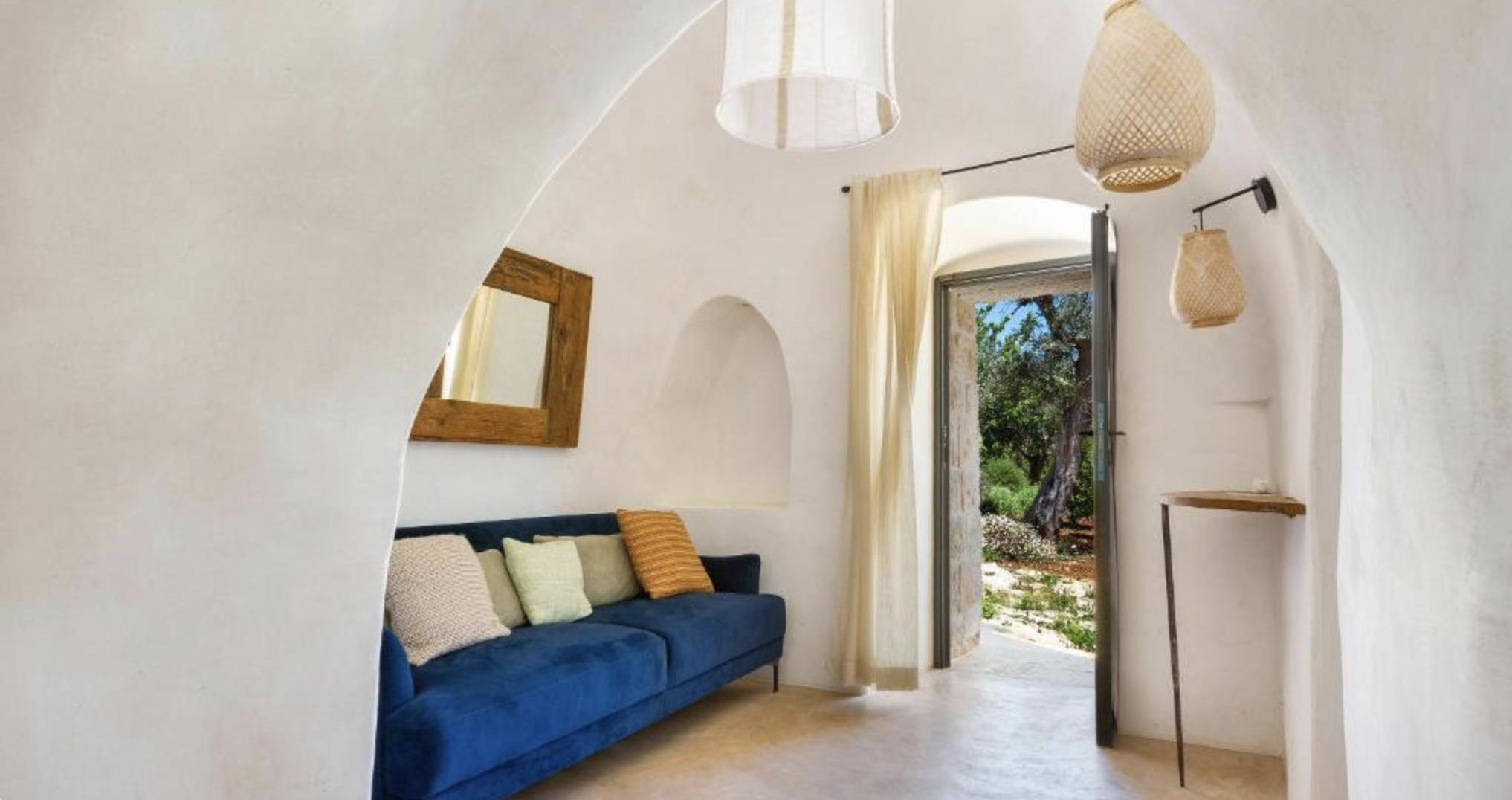 Intérieur d'une pièce avec un canapé bleu, coussins, murs blancs, une fenêtre avec vue sur un jardin, et des lampes en osier suspendues.