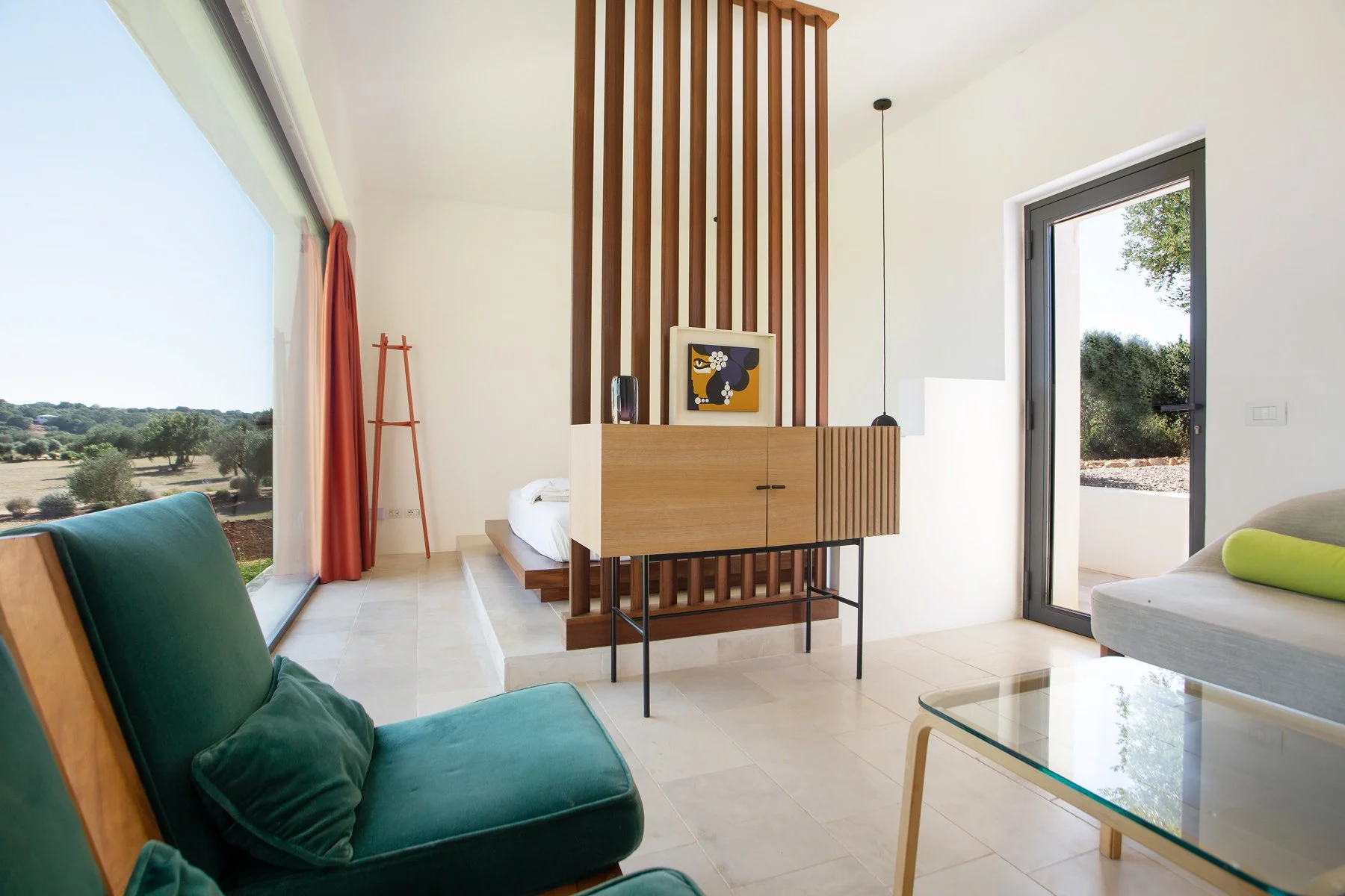 Salon lumineux avec grande fenêtre donnant sur un paysage naturel, canapé vert foncé avec coussins, table en verre, porte-fenêtre au fond, séparation en bois avec décoration moderne, ambiance claire et moderne.