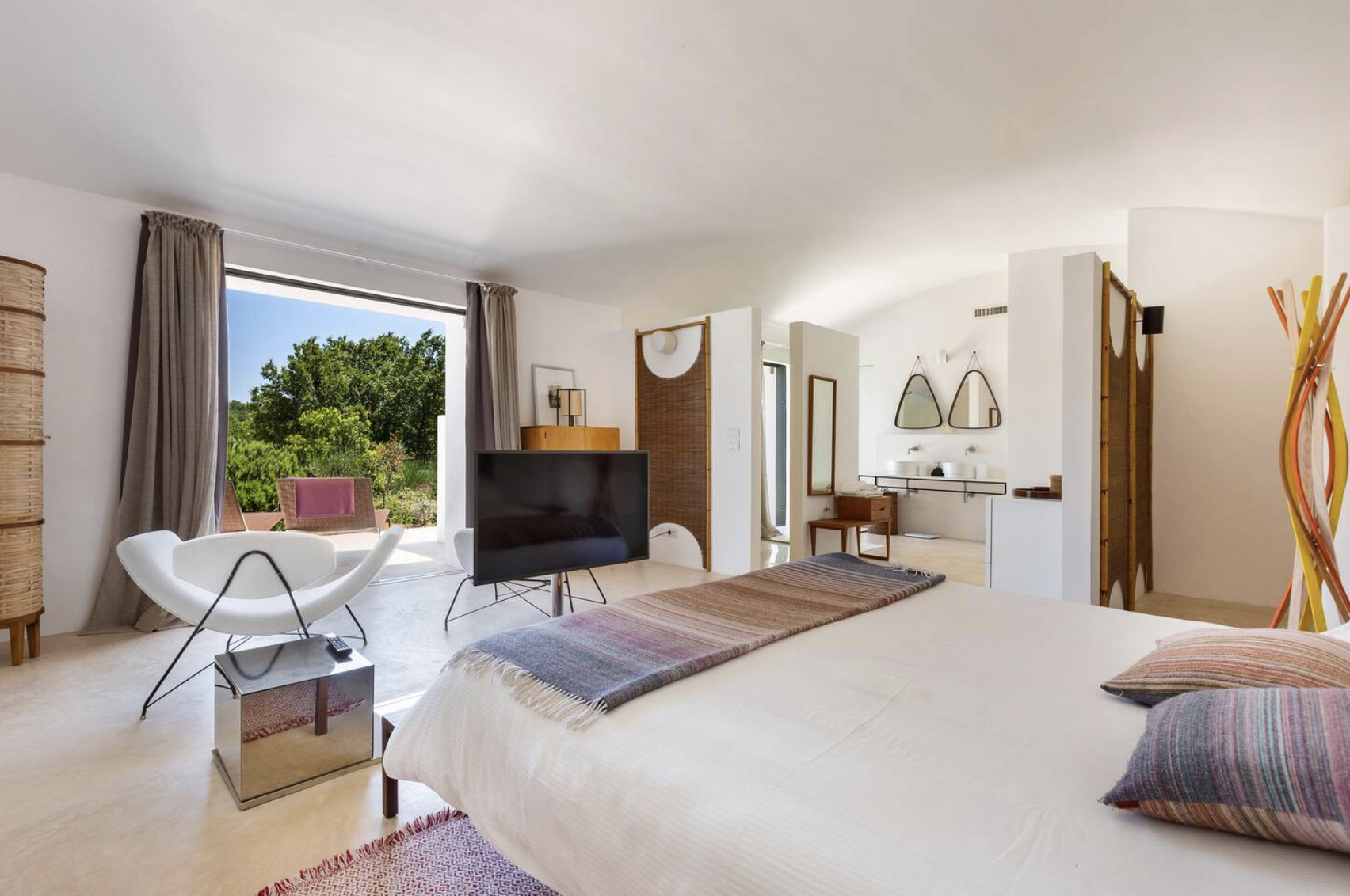 Chambre moderne avec vue sur la nature, lit avec une couverture à rayures, télévision, fauteuil blanc, autres meubles en bois, miroir, décoration minimaliste, grande fenêtre laissant entrer la lumière naturelle.