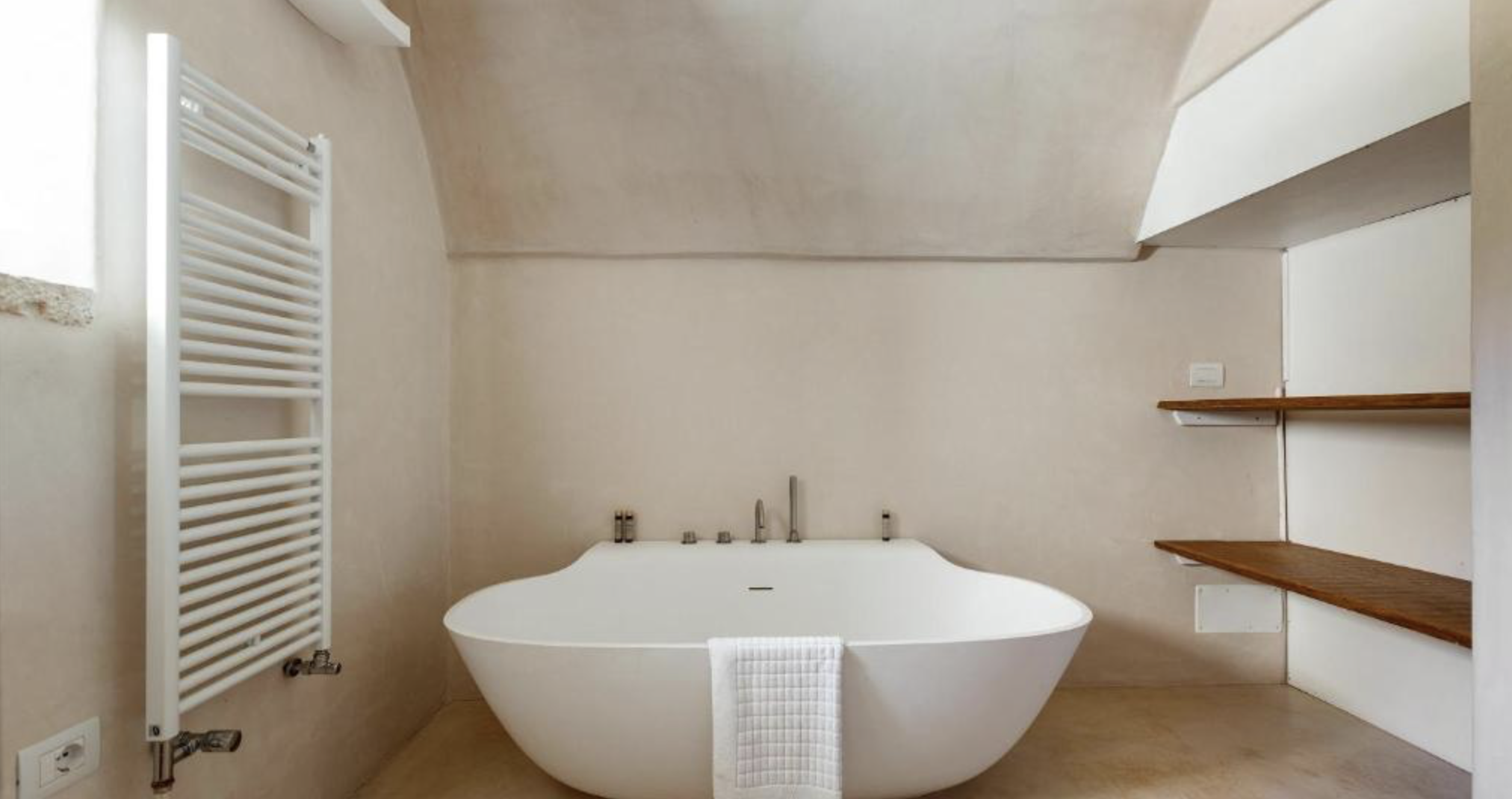 Baignoire blanche dans une salle de bain avec des murs beige, étagères en bois, radiateur blanc et prises électriques.