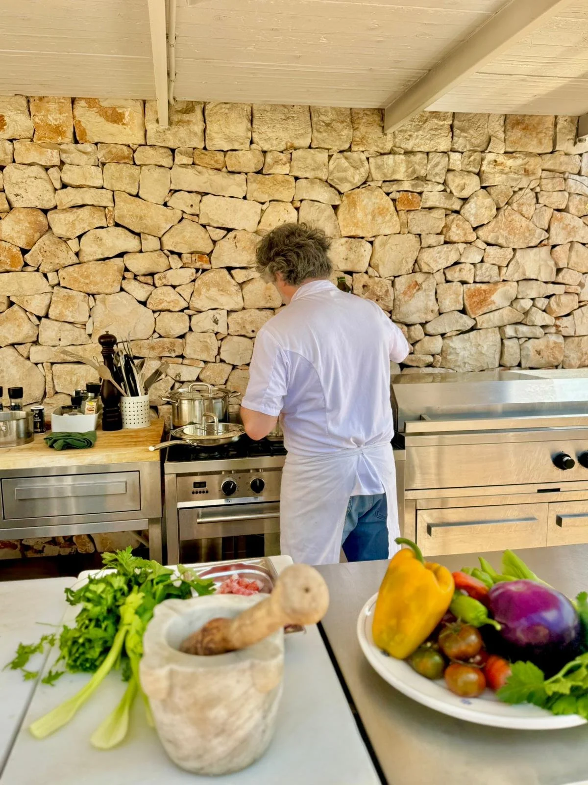 Un chef en blanc prépare des plats dans une cuisine avec un mur en pierres naturelles derrière lui, des légumes frais dans un bol en avant-plan.