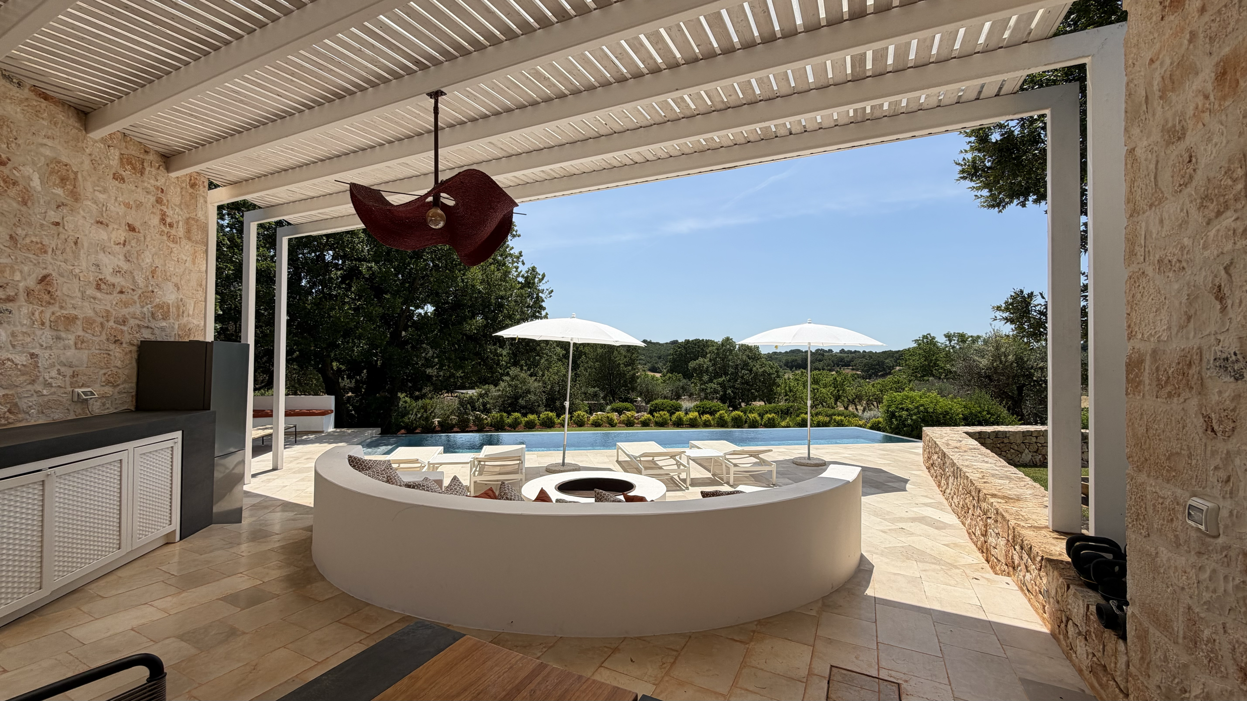 Vue depuis une terrasse couverte vers une piscine avec des parasols et des chaises longues, entourée de verdure et de collines en arrière-plan.