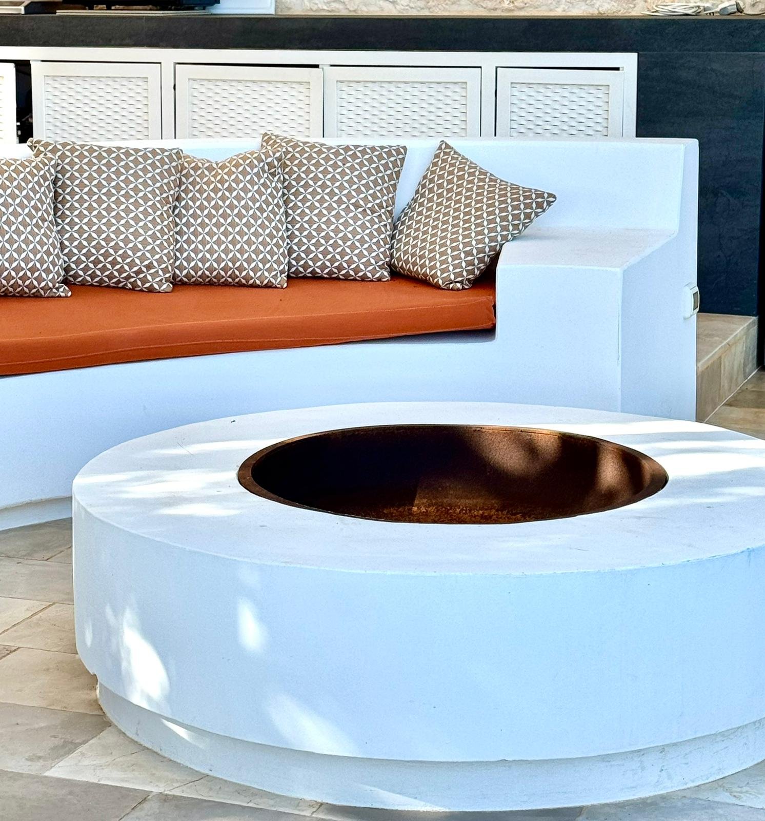 Un canapé blanc avec des coussins à motif géométrique beige et blanc, une table basse blanche ronde avec un trou central de couleur bronze ou cuivre, et un fond composé de panneaux blancs et d'un mur sombre.
