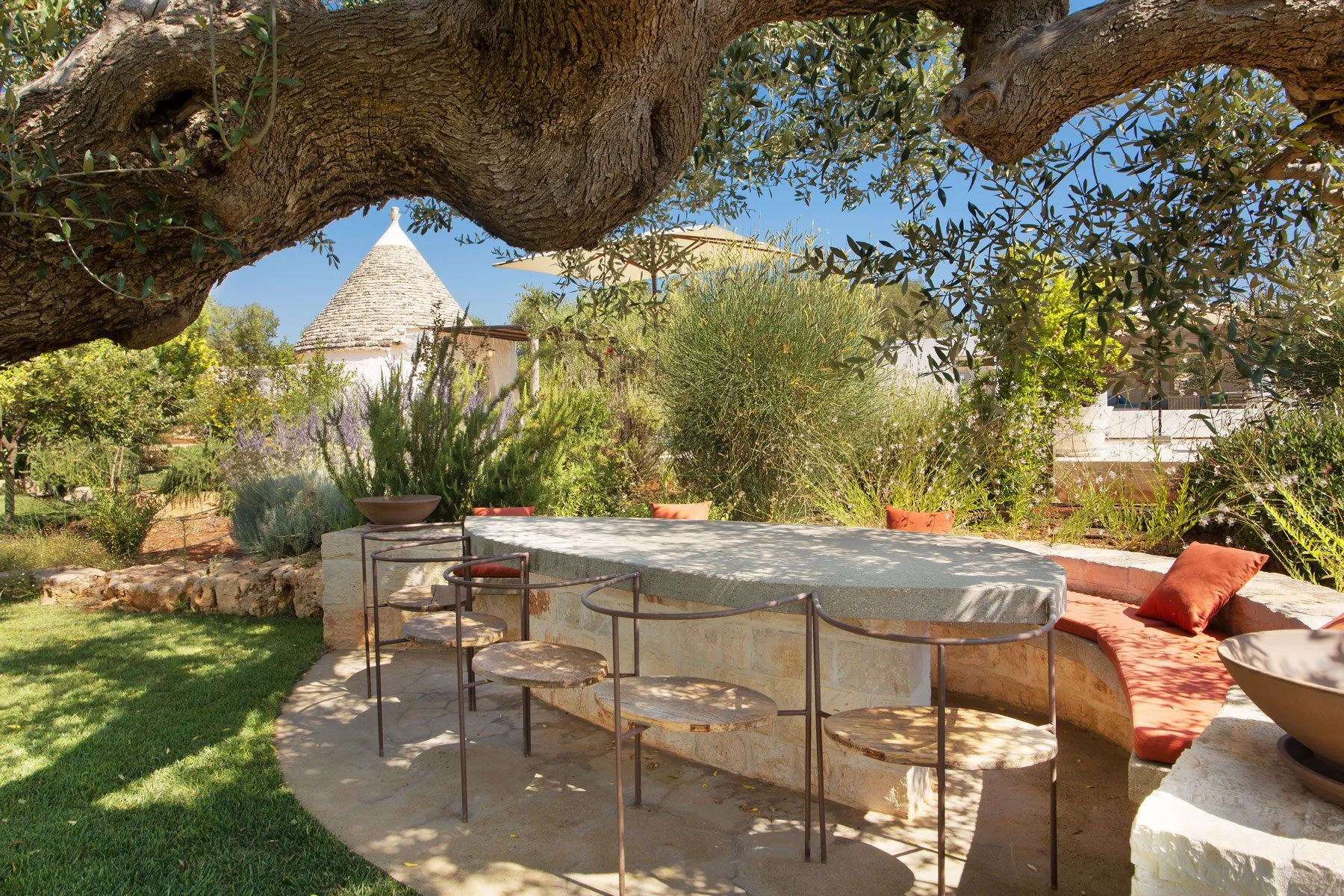 Un jardin méditerranéen avec une table ronde en pierre, des chaises en métal et des coussins orange, ombragé par un ancien olivier. En arrière-plan, des buissons, des plantes méditerranéennes et une maisonnette typique avec un toit en pierre.
