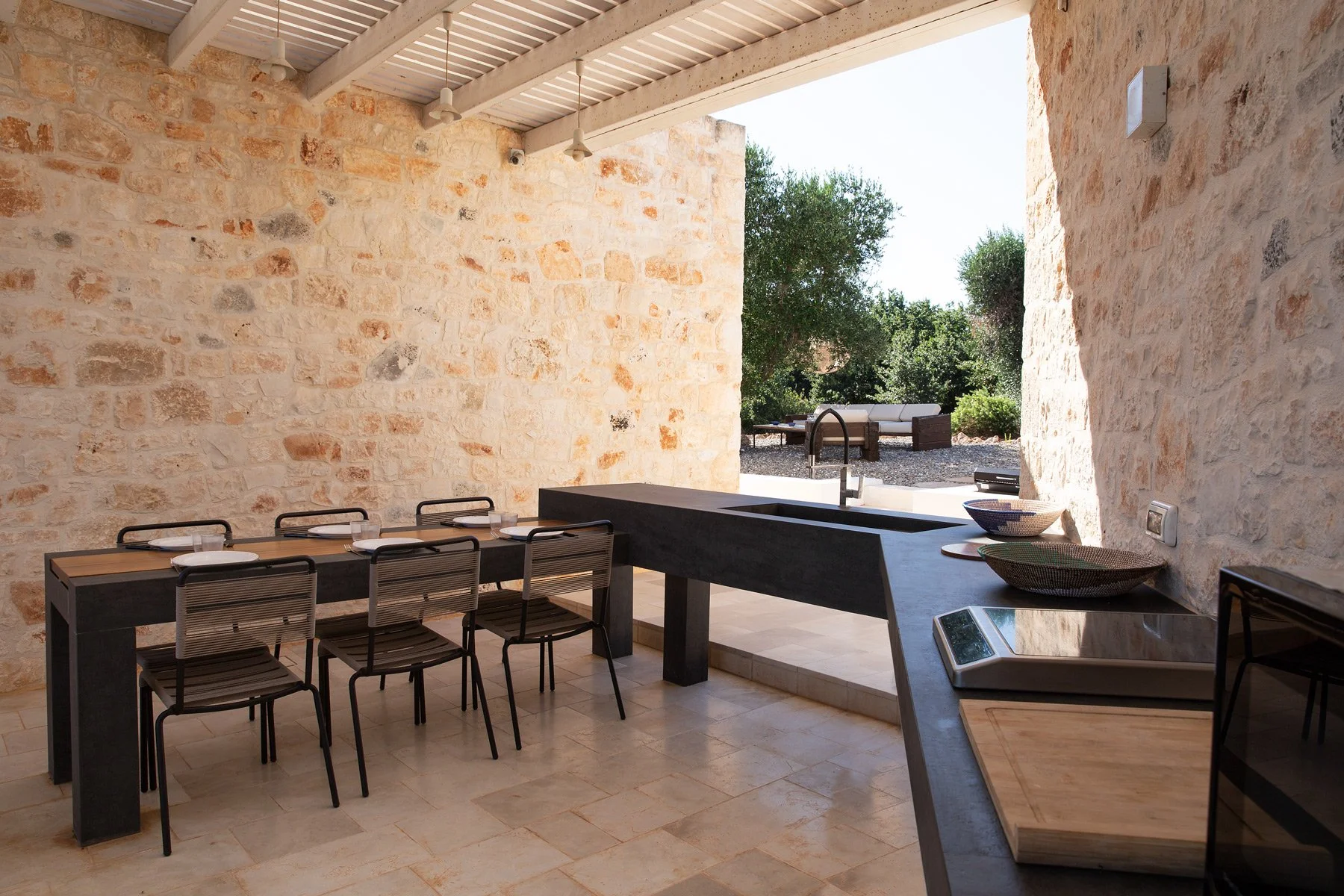 Intérieur d'une cuisine ou salle à manger avec murs en pierre, un plan de travail noir, une table avec des chaises et une vue vers l'extérieur avec des meubles de jardin et des arbres.