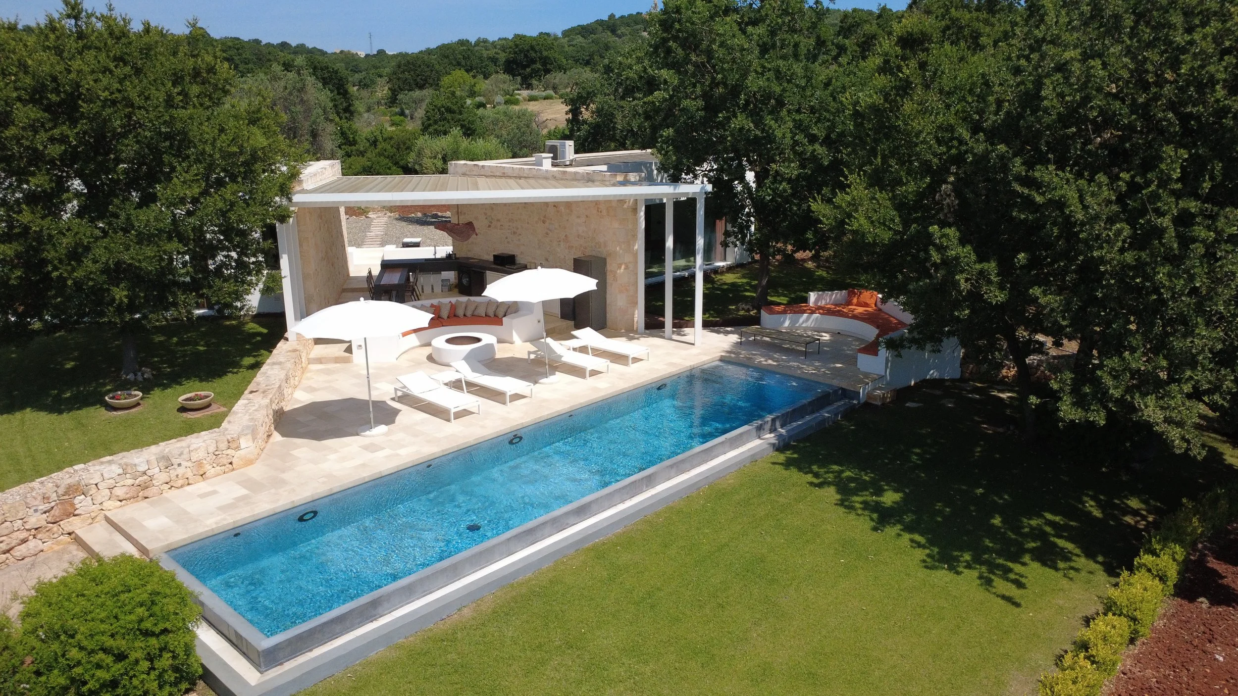 Piscine rectangulaire entourée d'une terrasse avec parasols et sièges, maison moderne en pierre avec espace extérieur, arbres et végétation environnante.