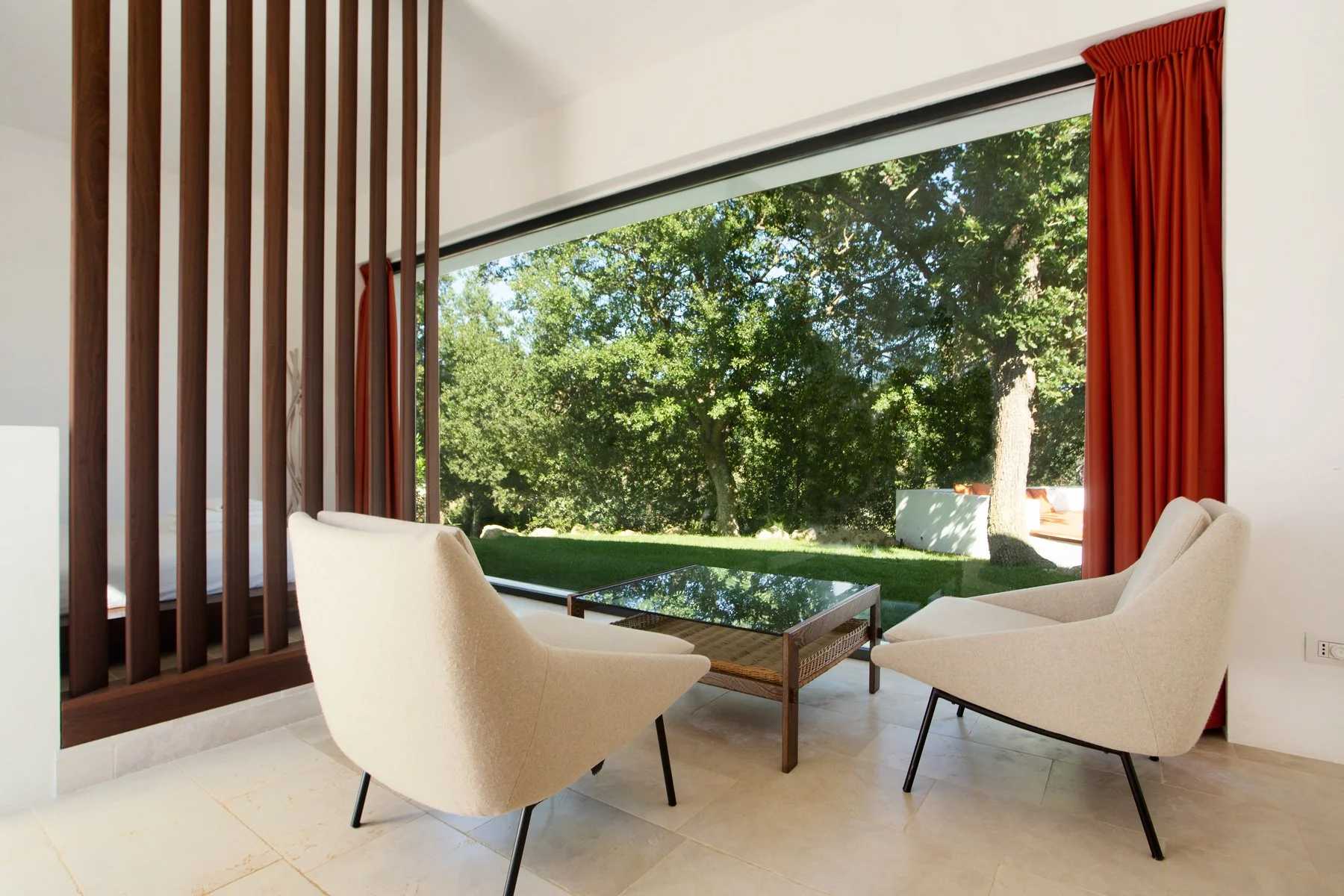 Salon moderne avec fauteuils blancs, table en verre et grande fenêtre donnant sur un jardin avec des arbres.