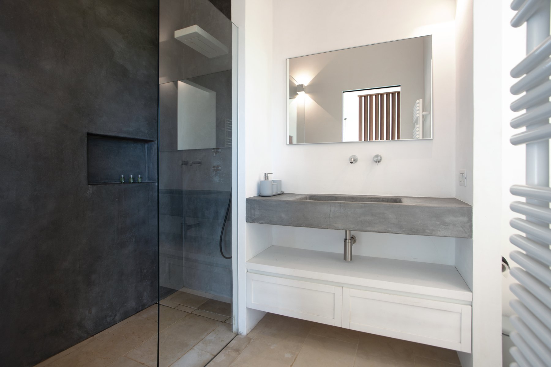 Salle de bain moderne avec un miroir rectangulaire au-dessus d'un lavabo en béton, avec un éclairage indirect et un radiateur à panneaux blancs à droite, et une cabine de douche vitrée avec un mur en pierre sombre à gauche.
