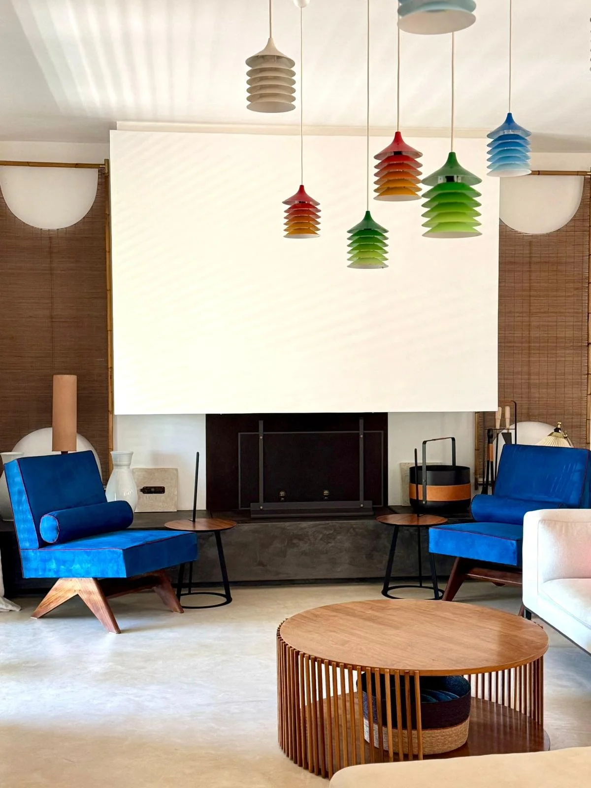 Salon moderne avec canapé blanc et fauteuils bleus, tables basses en bois, suspension colorée au plafond, cheminée encadrée en noir, décor en bois et accessoires divers.