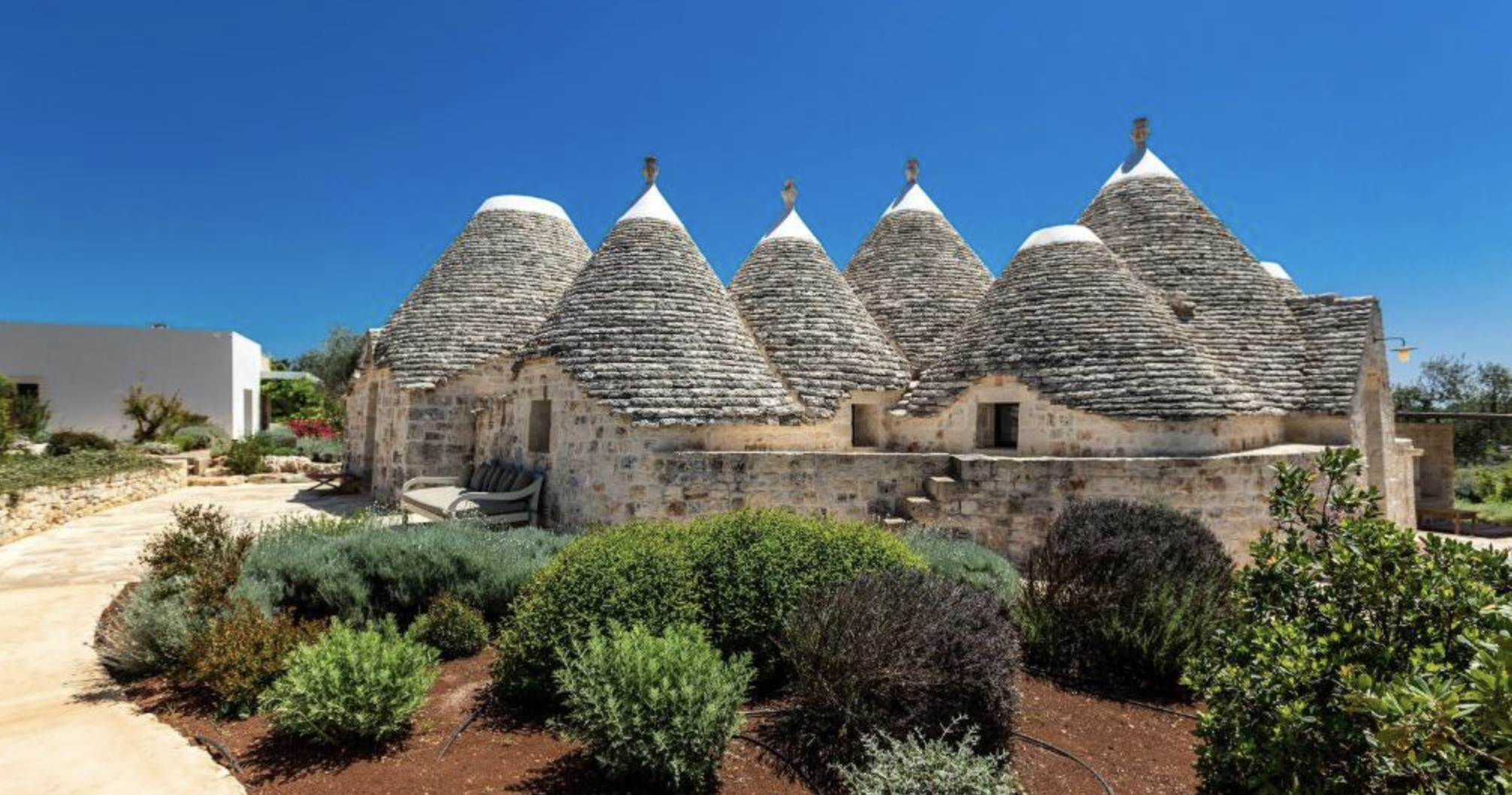 Une structure de style traditionnel italien faite de pierre, avec plusieurs toits coniques recouverts de neige, située dans un jardin bien entretenu sous un ciel bleu clair.