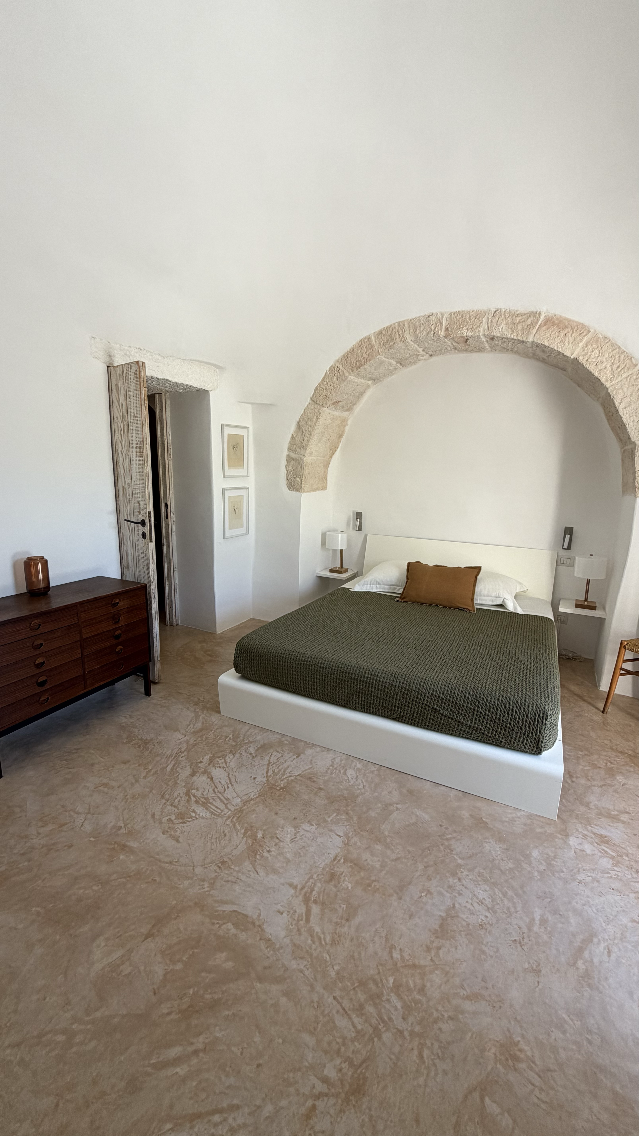 Chambre moderne avec un lit à cadre blanc, une tête de lit blanche, deux lampes de chevet, un mur en brique arquée, deux tableaux encadrés, un meuble en bois foncé, et un sol en pierre beige marbrée.