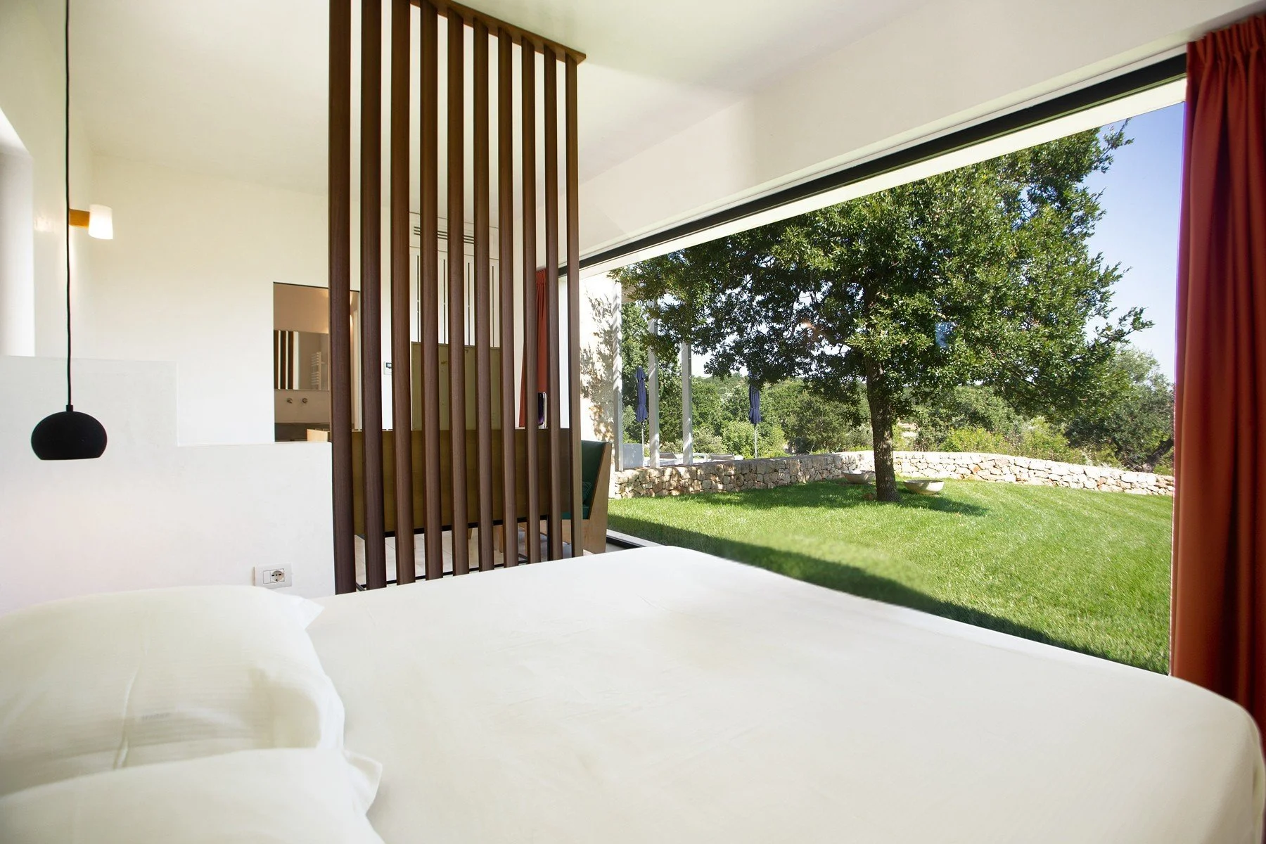 Vue depuis une chambre d'hôtel avec un lit blanc, une grande fenêtre laissant entrer la lumière naturelle, vue sur un jardin avec un arbre et une pelouse, avec un mur en pierre en arrière-plan.