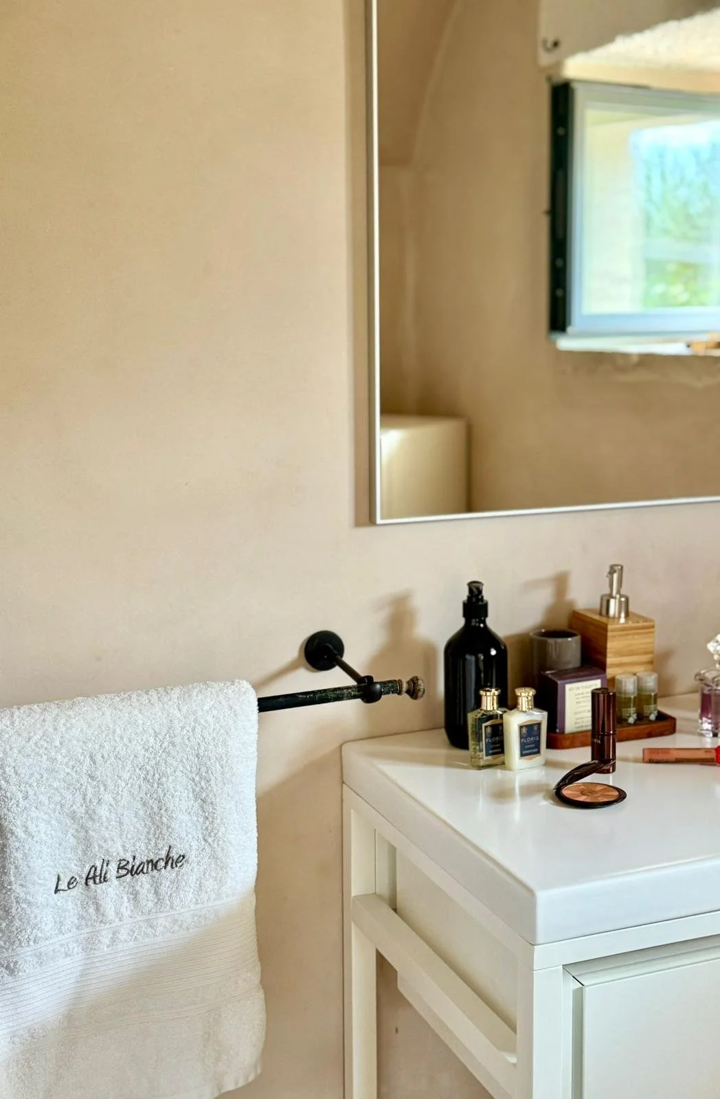 Un miroir au-dessus d'un lavabo blanc avec divers produits de toilette, une serviette rose avec l'inscription "Le All Blanche" suspendue à une barre noire, une fenêtre laissant entrer de la lumière naturelle, dans un intérieur de salle de bain élégan