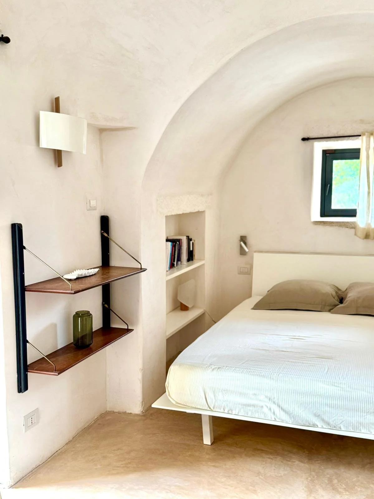 Chambre avec lit blanc, deux oreillers gris, petite étagère intégrée avec livres, fenêtre avec rideau blanc, mur blanc avec alcôve, et lampe murale.
