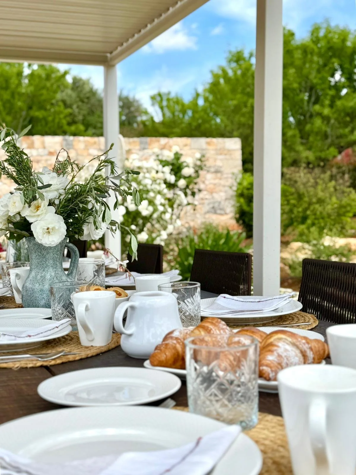 Table de petit-déjeuner en plein air avec croissants, tasses, verres et fleurs, sous une pergola avec vue sur un jardin verdoyant.