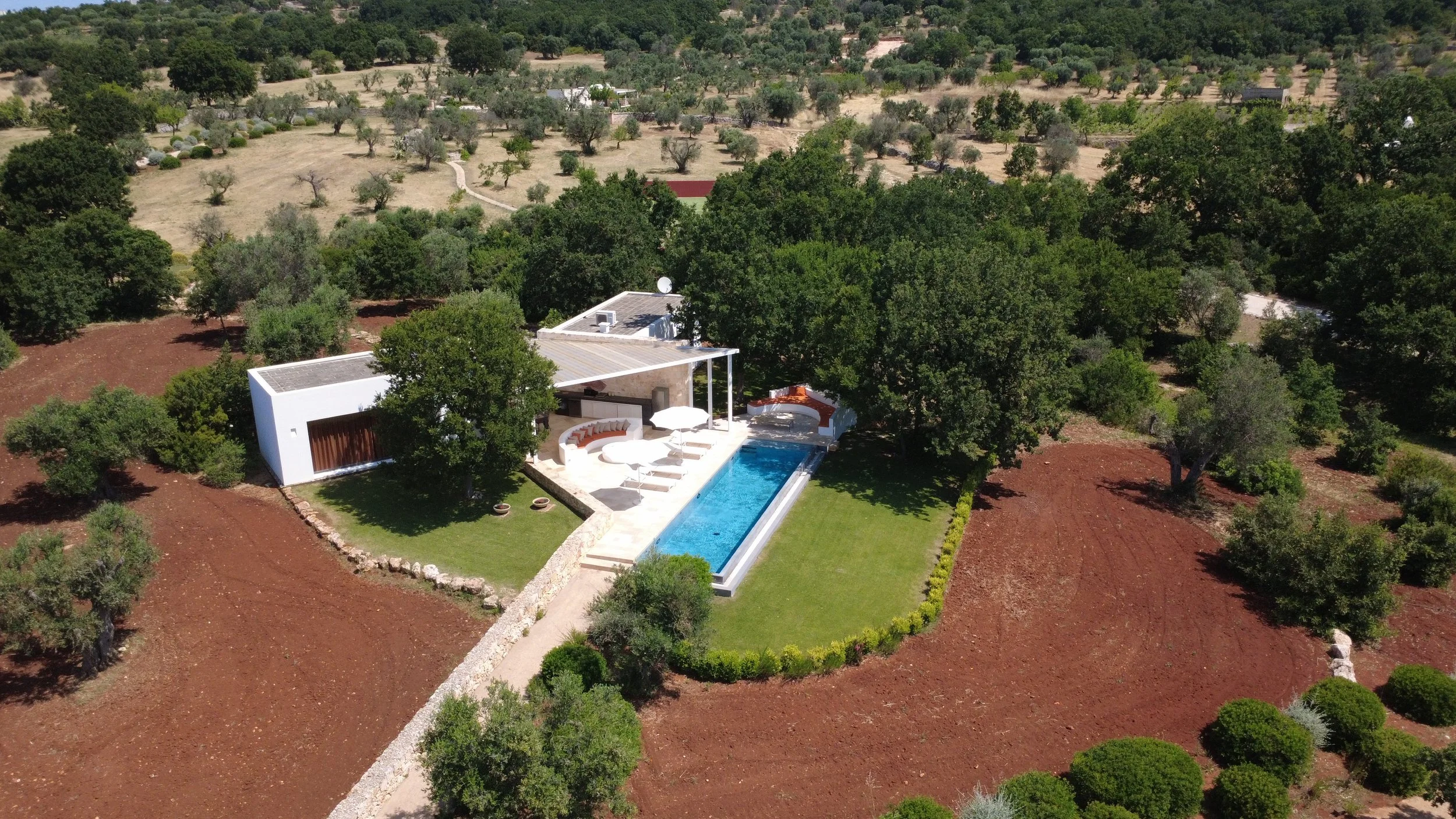 Vue aérienne d'une maison moderne avec une piscine, entourée de pelouse et d'arbres, dans une région rurale avec des oliviers et des collines arides au loin.