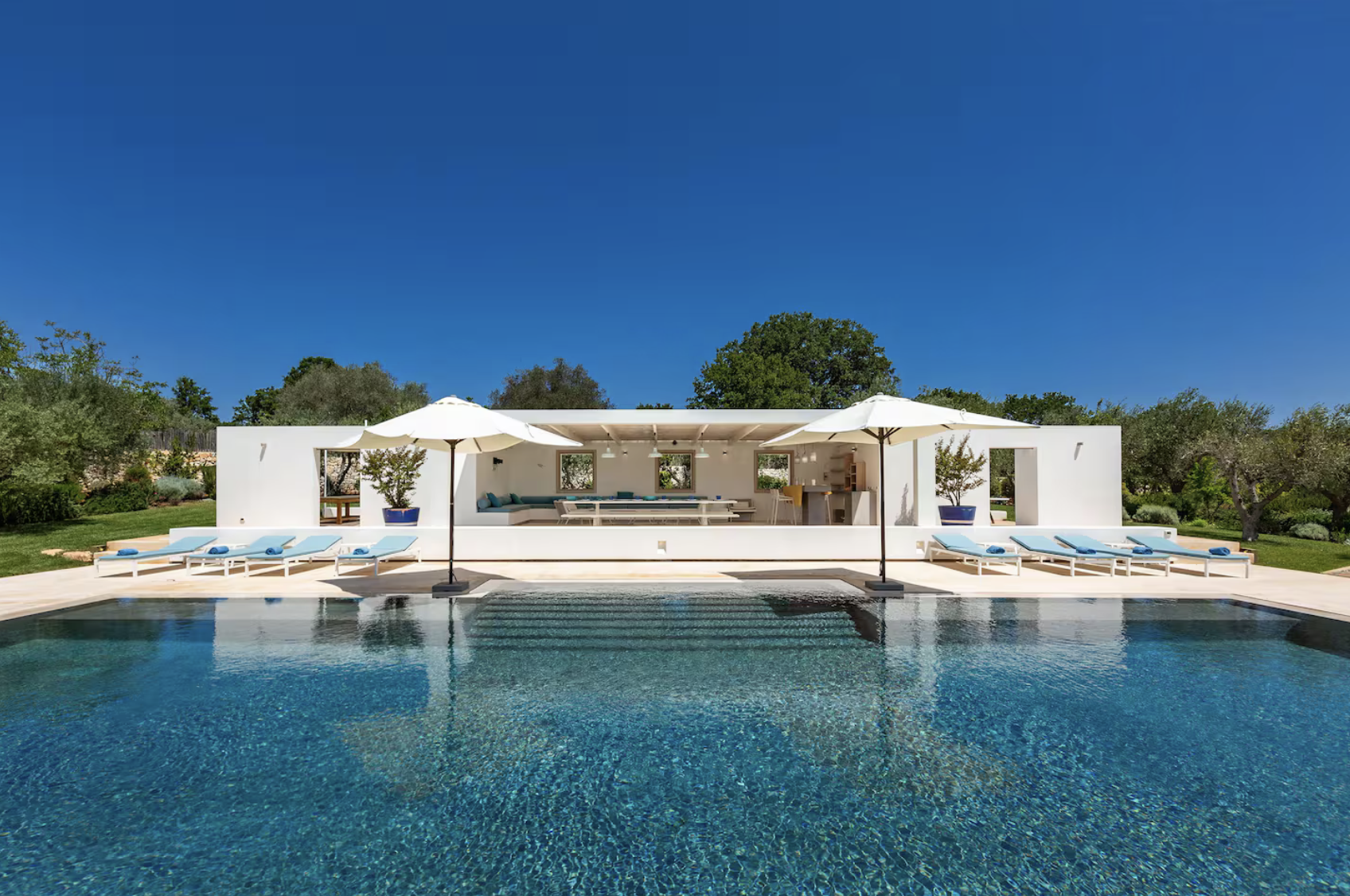 Une piscine devant une maison moderne blanche avec un espace salon à l'intérieur et deux parasols, entourée de végétation et sous un ciel bleu.