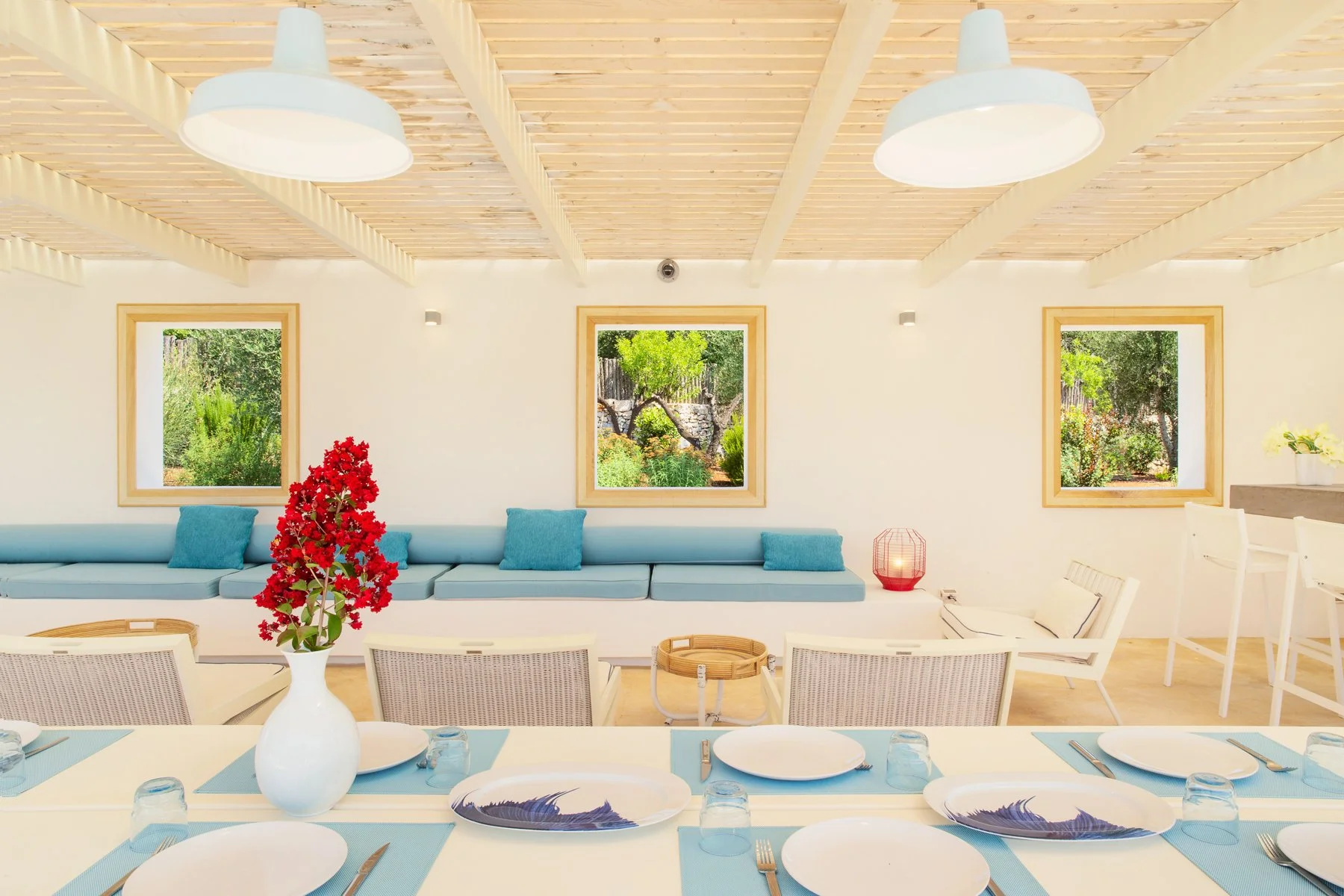 Salle à manger lumineuse avec fenêtres donnant sur un jardin, décorée dans un style minimaliste avec des chaises blanches, des assiettes blanches, une nappe blanche avec des sets bleus, et une vase avec des fleurs rouges sur la table.