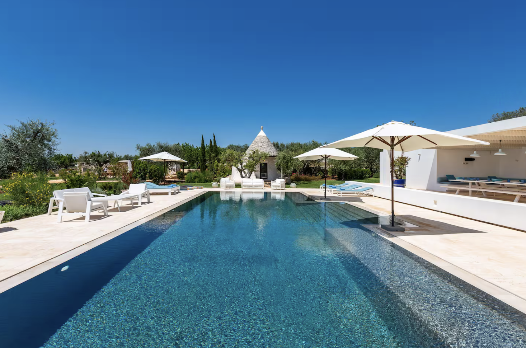Piscine extérieure avec parasols blancs et mobilier de jardin blanc, jardin fleuri en arrière-plan, ciel bleu clair, maison moderne avec espace détente.