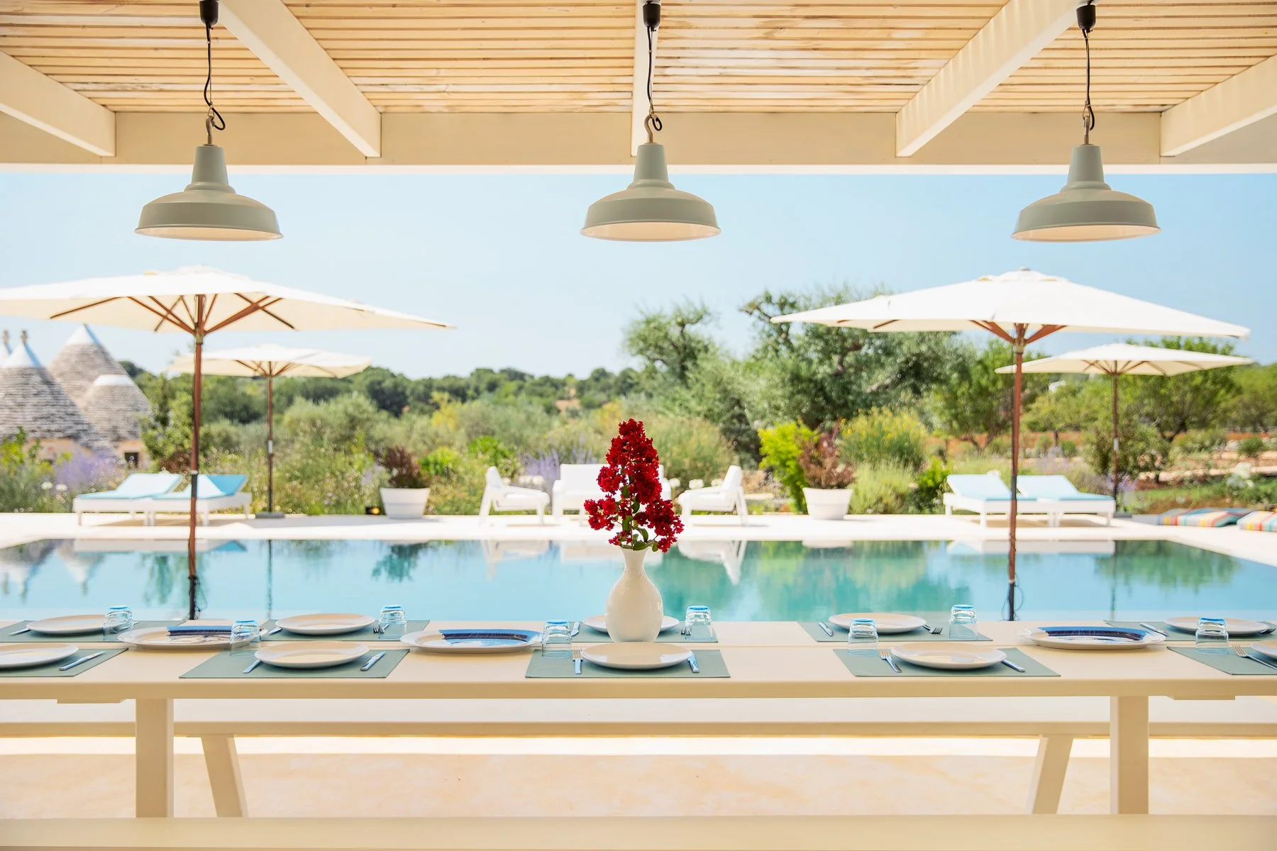 Table de repas avec assiettes, couverts, verres et une vase avec des fleurs rouges, vue sur une piscine et des parasols en plein air dans un jardin ensoleillé.