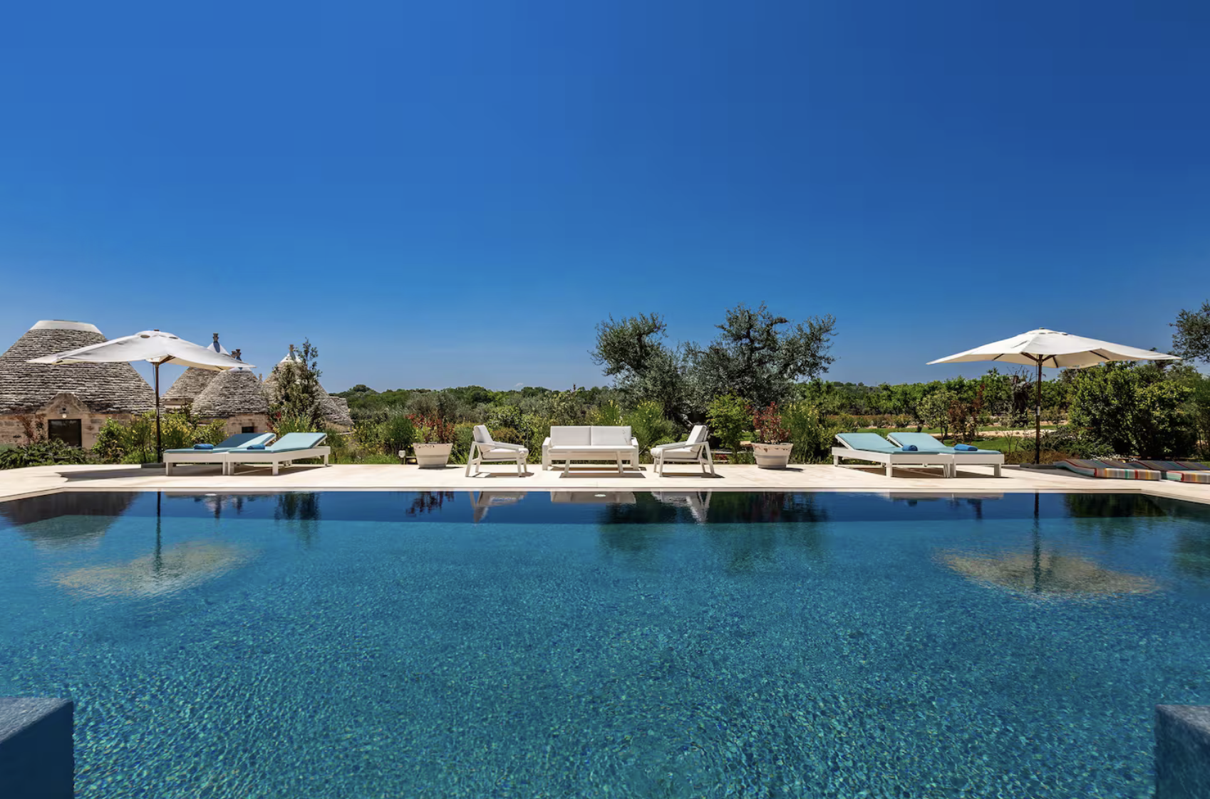 Piscine extérieure avec transats et parasols blanc, bâtiments traditionnels en pierre en arrière-plan, sous un ciel bleu clair.
