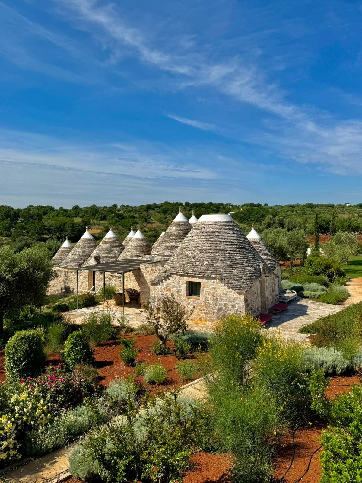 Un groupe de maisons en pierre avec des toits coniques en forme de chapiteau, dans un paysage verdoyant avec des arbres et un ciel bleu.