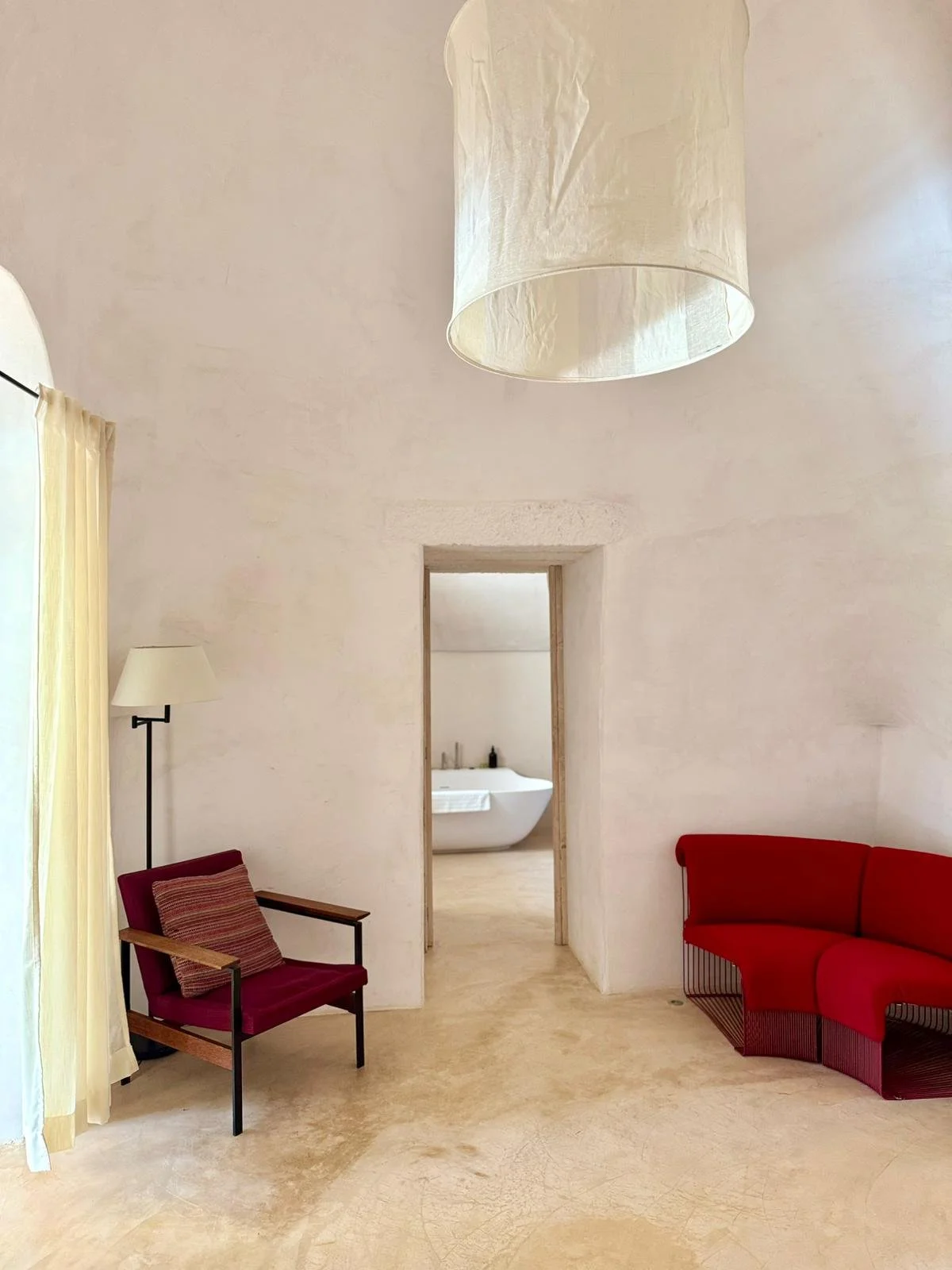 Une pièce avec des murs blancs texturés, un tapis de sol beige, une porte menant à une salle de bain avec une baignoire blanche. Sur la gauche, un fauteuil en bois avec un coussin rayé rouge et beige, une lampe à pied blanche à côté. Sur la droite, u