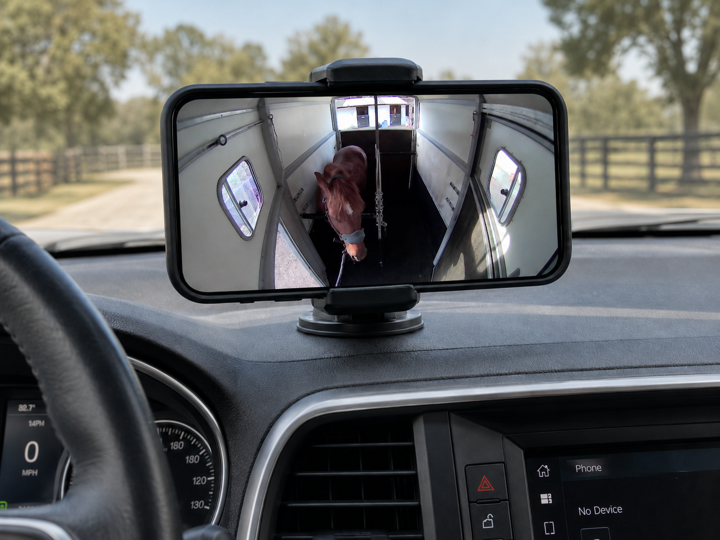 A view from inside a vehicle showing its dashboard, with a mobile device mounted on the dashboard displaying a backing camera view of a horse in a trailer.