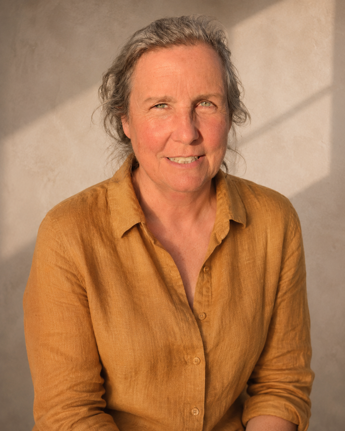 A middle-aged woman with wavy gray hair, smiling, wearing a brown button-up shirt, standing in front of a plain wall with shadow patterns.