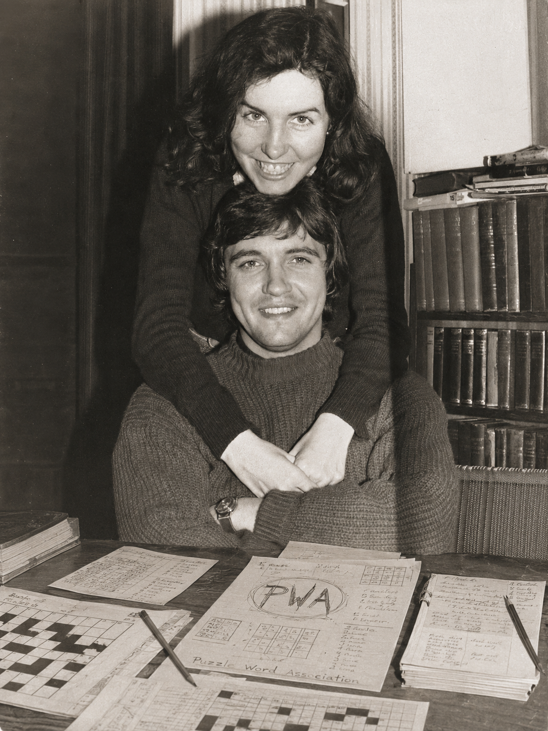 A woman with dark, curly hair leaning over a young man with straight hair, both smiling, surrounded by papers, books, and crossword puzzles in a room with wood-paneled walls.
