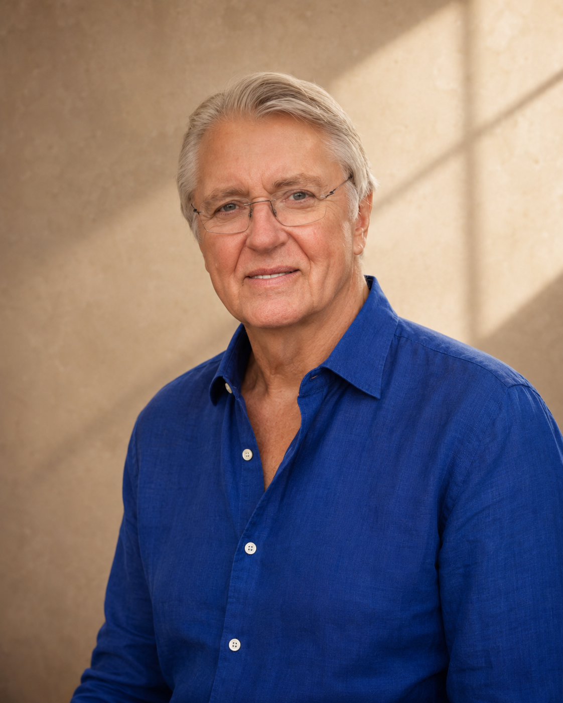 A middle-aged man with gray hair, wearing glasses and a blue button-up shirt, smiling softly in front of a beige background with sunlight shadows.