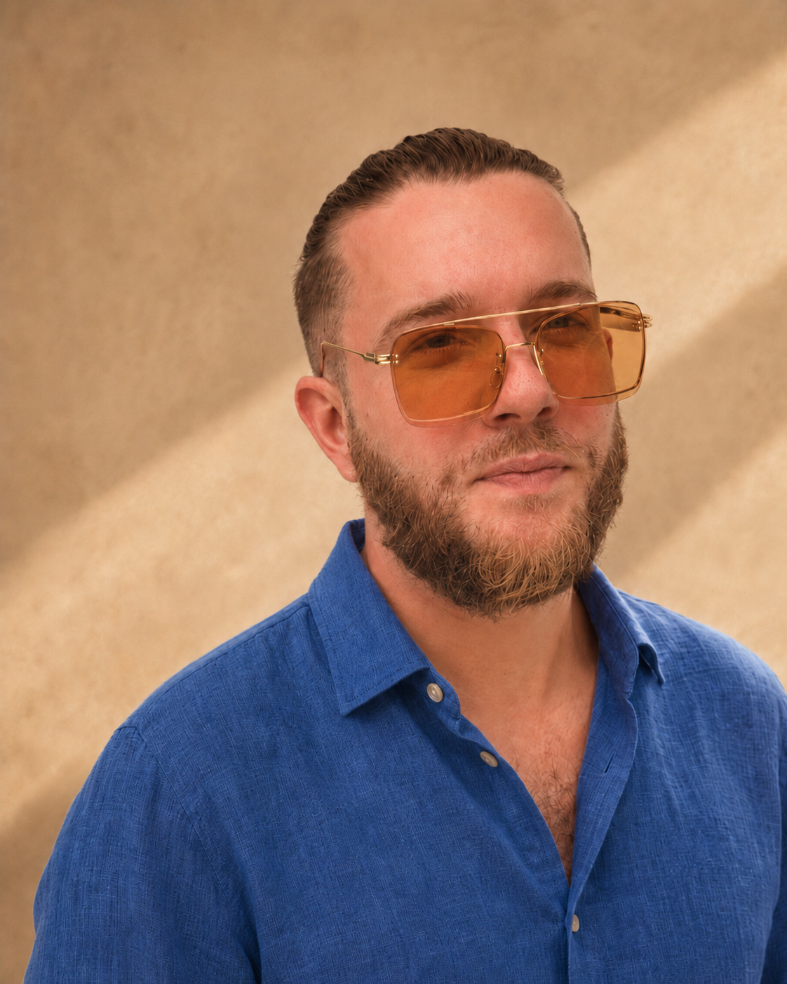 A man with a beard is wearing orange-tinted sunglasses and a blue collared shirt, standing against a gold and beige background.