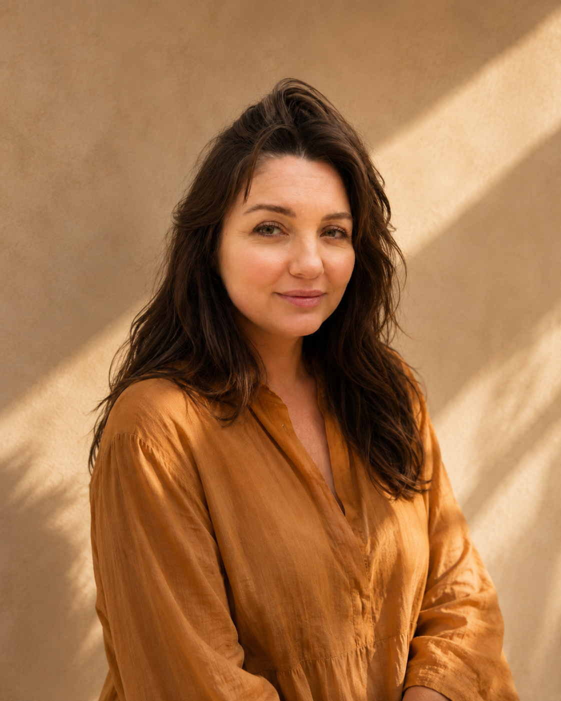 A woman with long, wavy brown hair smiling, wearing an orange-brown satin blouse, standing against a beige background with sunlight creating diagonal shadow lines.