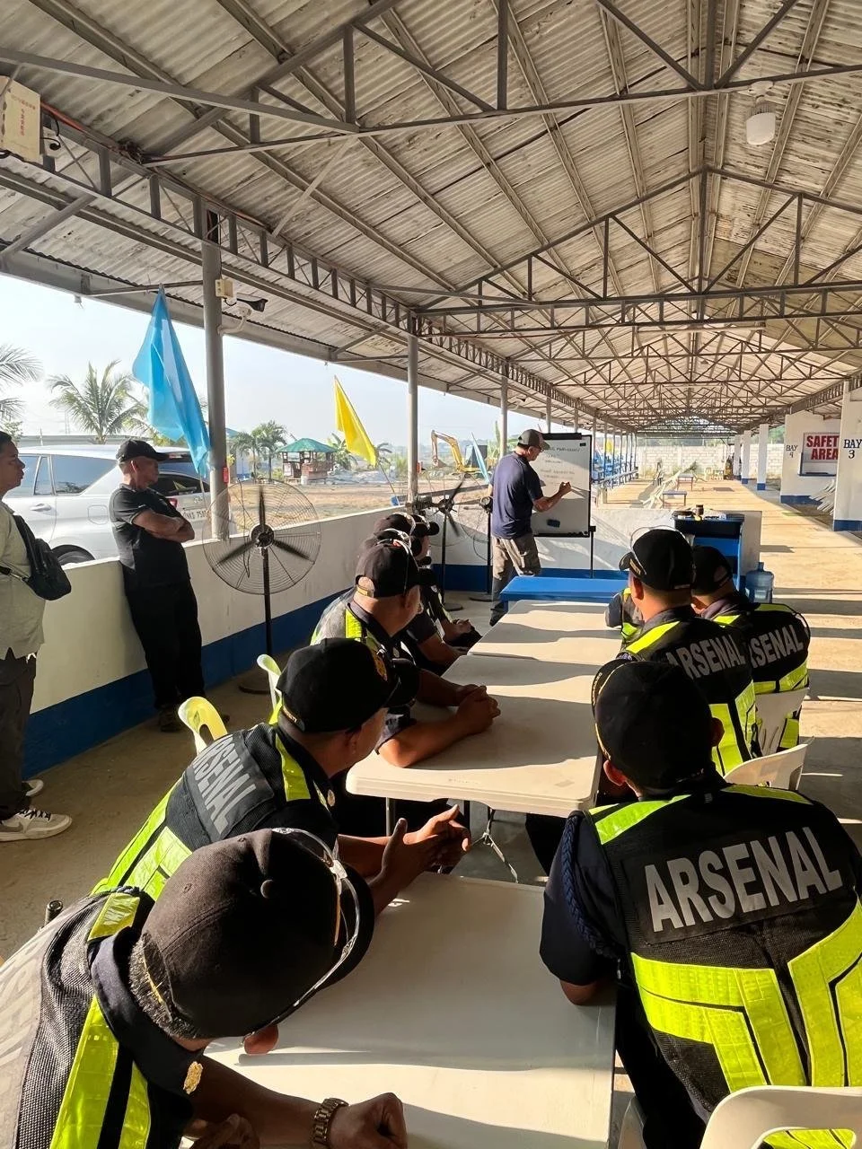 Police officers sitting at a long table inside a covered area, listening to a man at a whiteboard, with people standing nearby and flags hanging from the roof, possibly during a briefing or training session.