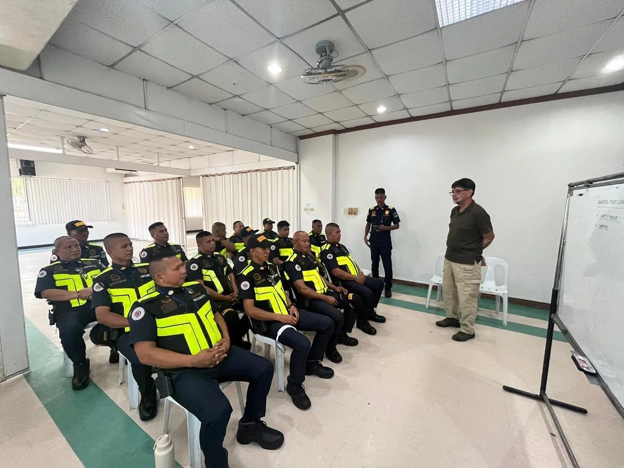 Police officers in uniform attending a training or briefing in a classroom setting, seated and listening to an instructor standing in front of them near a whiteboard.