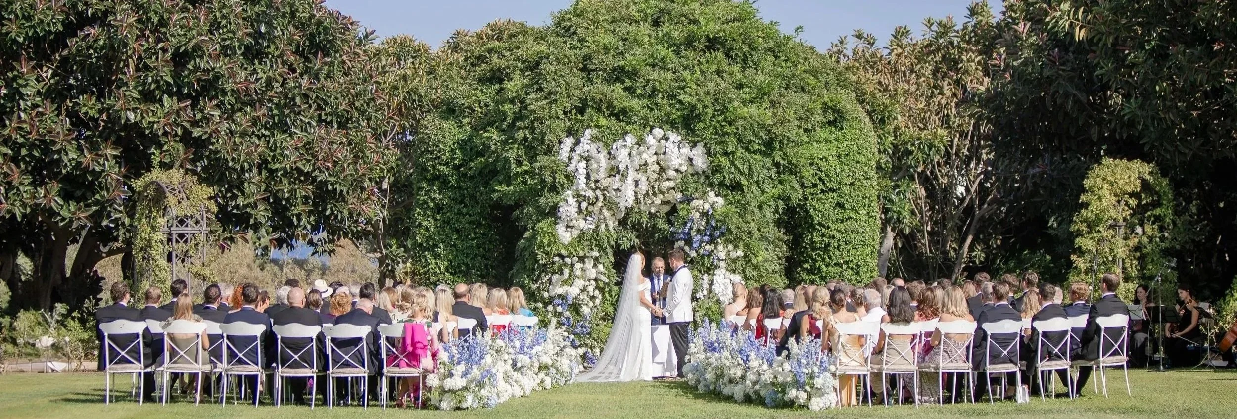 Outdoor wedding ceremony with bride and groom exchanging vows in front of an arch decorated with white flowers, surrounded by seated guests, with lush green trees in the background.
