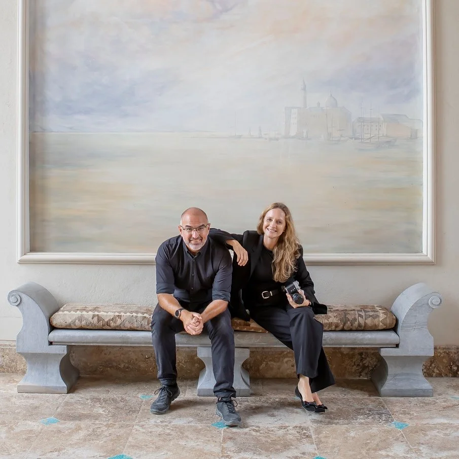 A man and woman sitting on a decorative stone bench inside a room, with a large abstract painting of a harbor scene hanging on the wall behind them. The woman is holding a camera, and both are smiling.